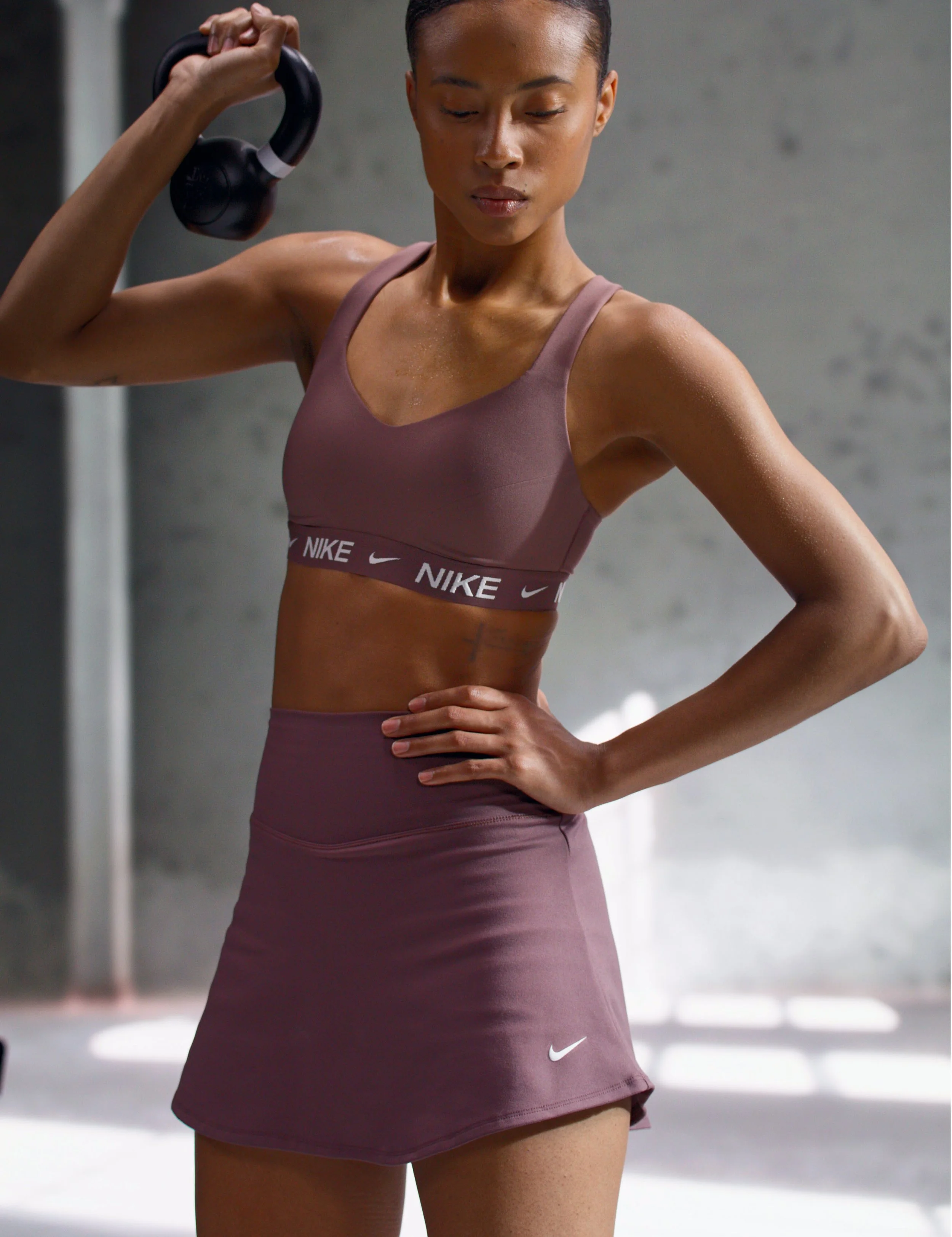 A woman in a pink Nike sports bra and matching skirt holding a black kettlebell on her shoulder, standing in a gym.