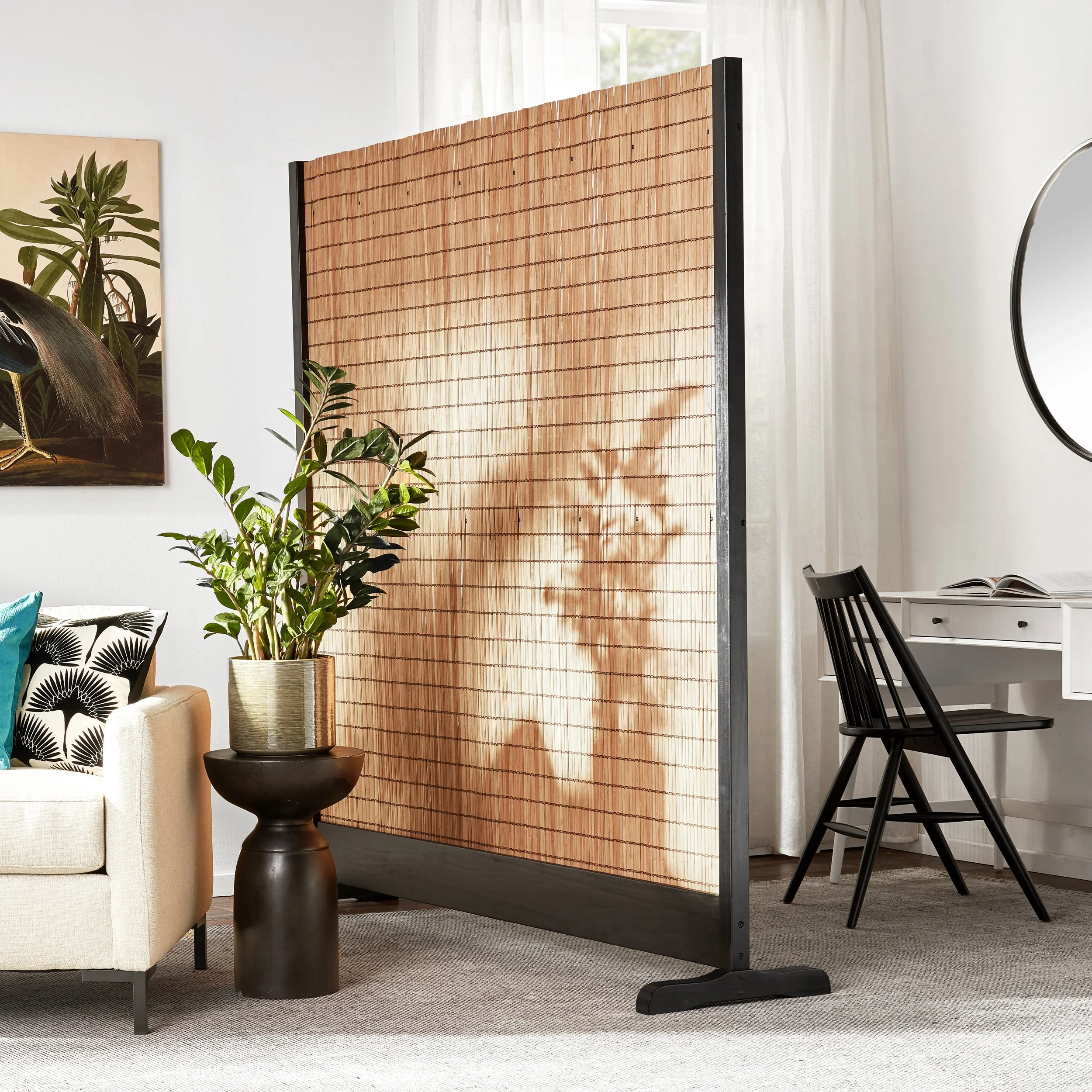 Room divider in front of a window with sheer curtains, a potted plant on a black pedestal, a black chair, and a white desk with open magazine in a bright, modern living space.