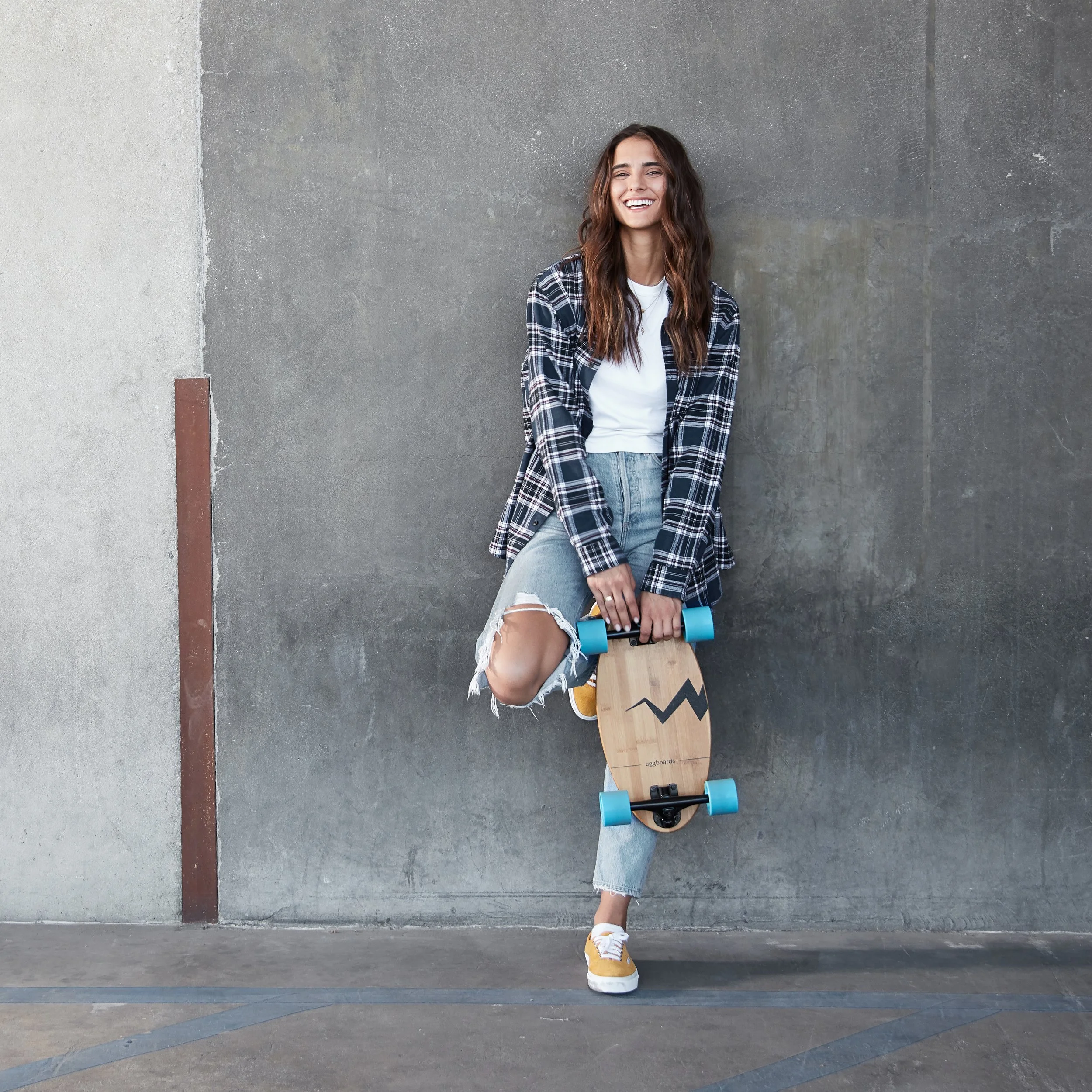 A young woman with long brown hair, wearing a plaid shirt, white t-shirt, ripped jeans, and yellow sneakers, smiling while holding a skateboard with blue wheels and a wooden deck, standing against a gray concrete wall.