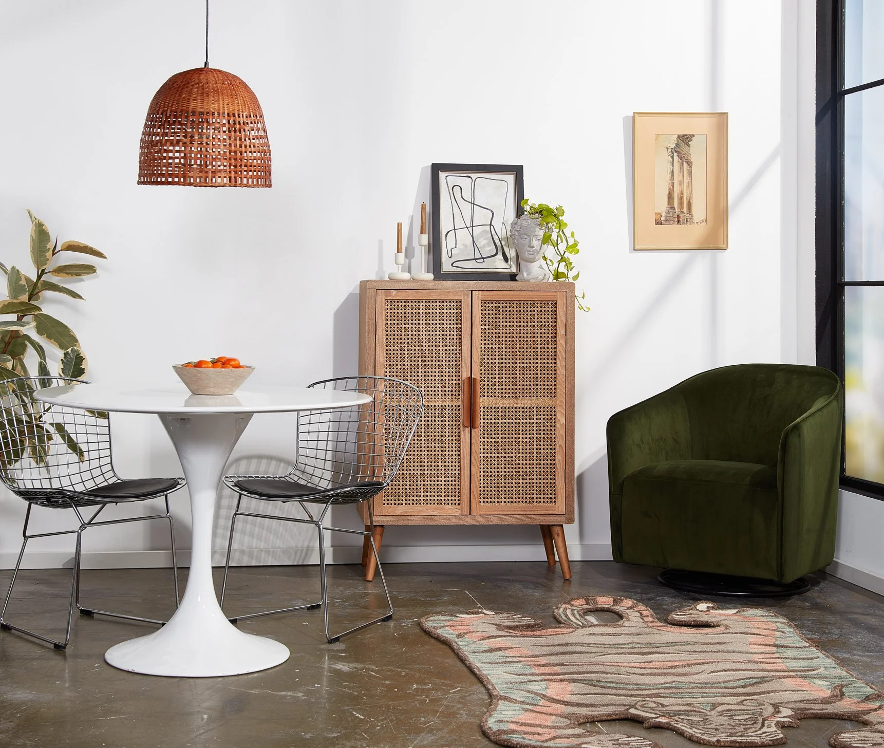 Interior of a modern living room with a white wall, wooden cabinet, round white dining table, black wire chairs, green velvet armchair, hanging wicker lamp, framed art, and a small rug on the wooden floor.
