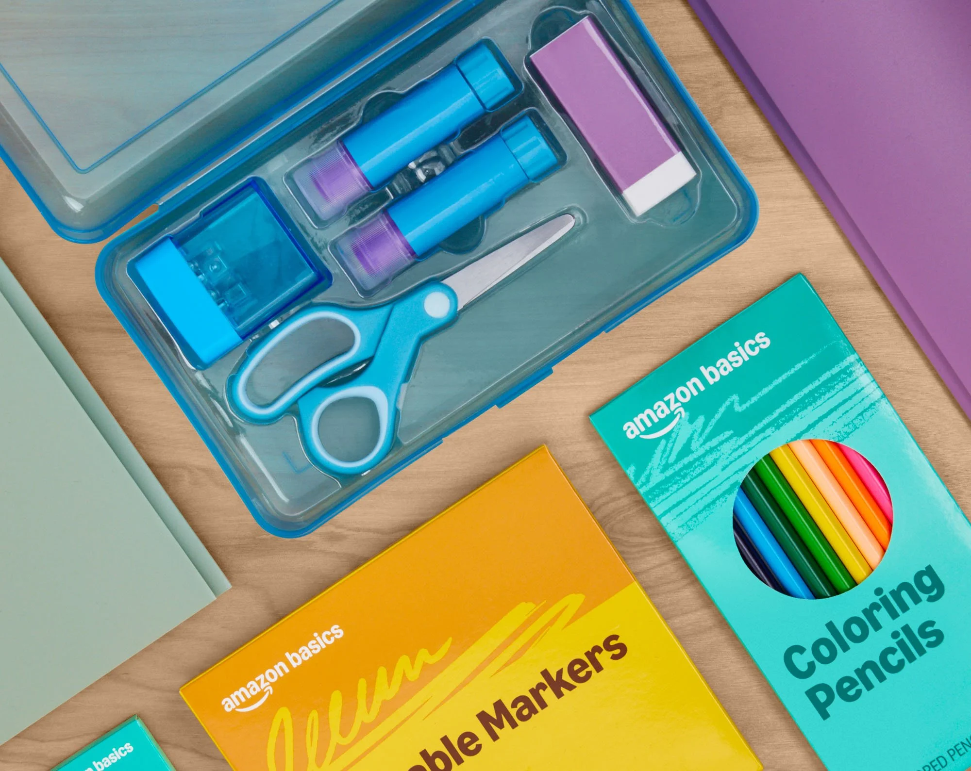 School supplies on a wooden surface, including a plastic case with scissors, glue sticks, and an eraser, alongside boxes of colored pencils and markers.