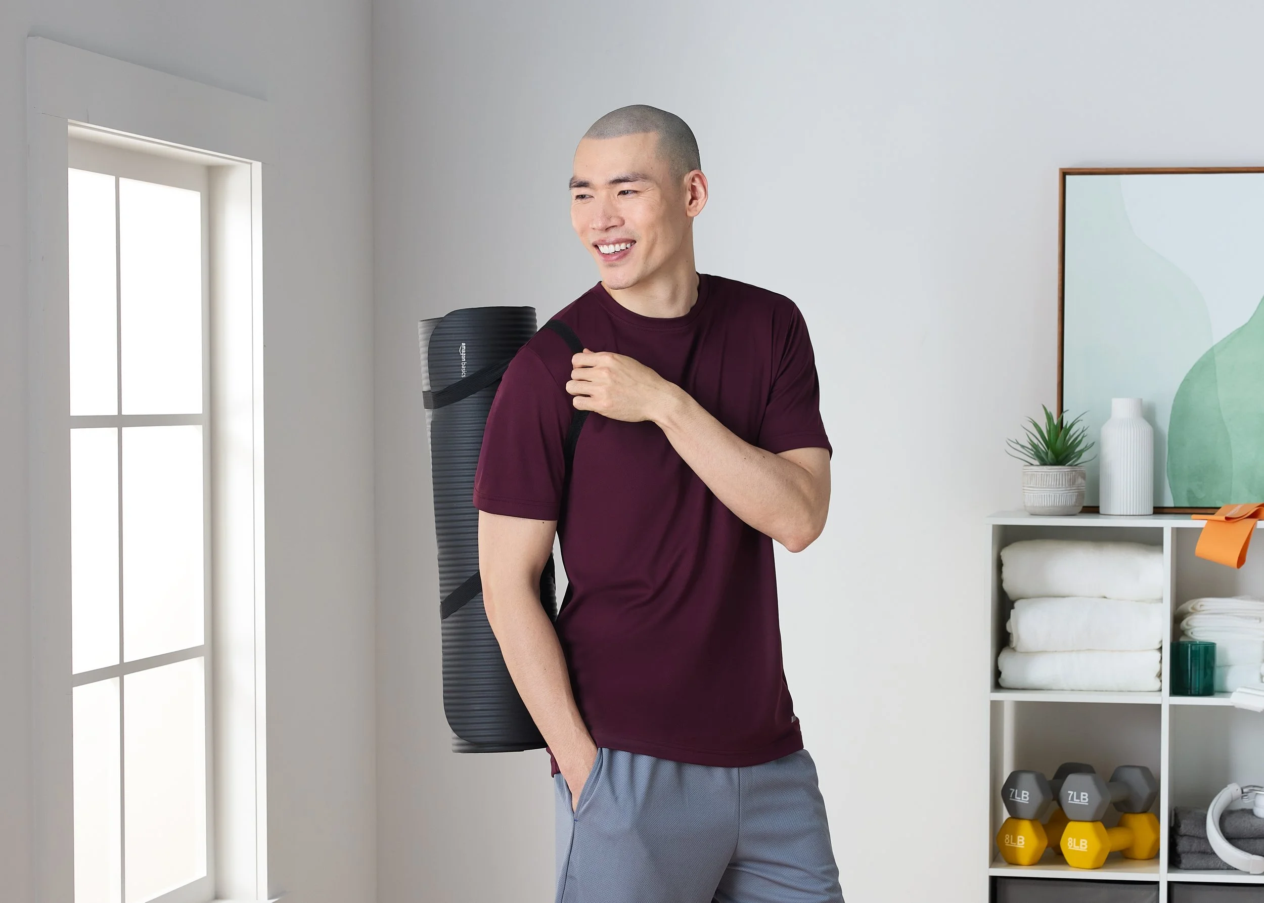 Young man with a shaved head, wearing a maroon shirt and gray shorts, carrying a black exercise mat on his shoulder, standing in a bright room with gym equipment and decor.