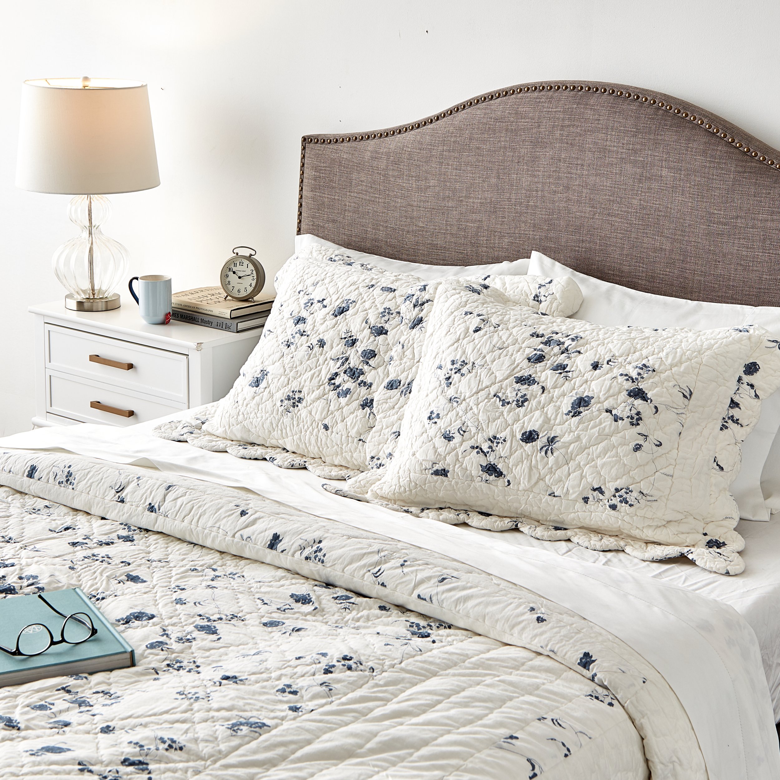 A bedroom with a bed that has a brown upholstered headboard, white and floral bedding, and matching pillows. A white nightstand with a lamp, clock, mug, and books sits beside the bed. A pair of glasses and a closed book are on the bed.