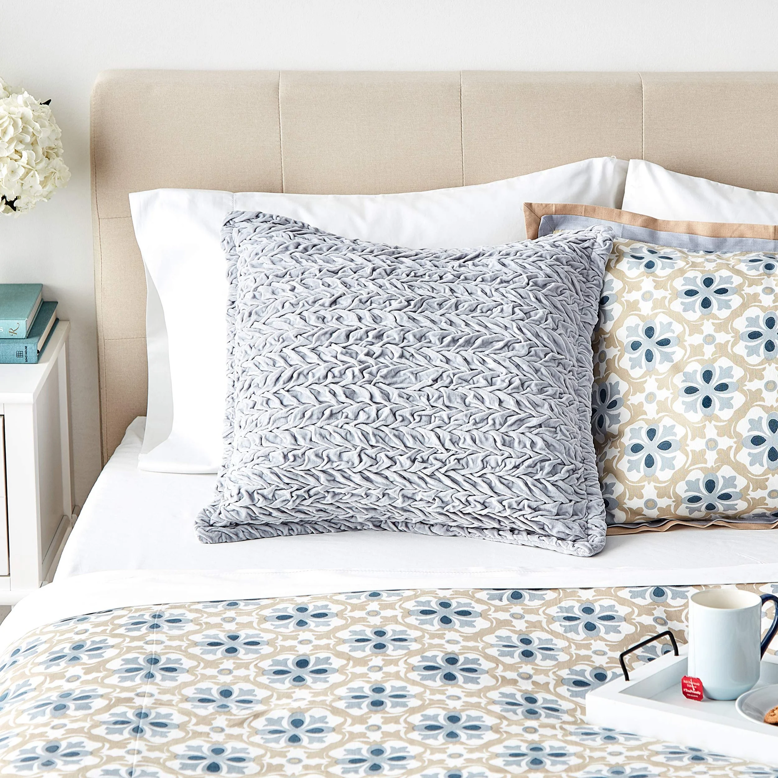 A neatly made bed with decorative pillows, a beige headboard, a white nightstand with books and a bouquet of white flowers, and a tray with a mug and tea bag on a patterned bedspread.