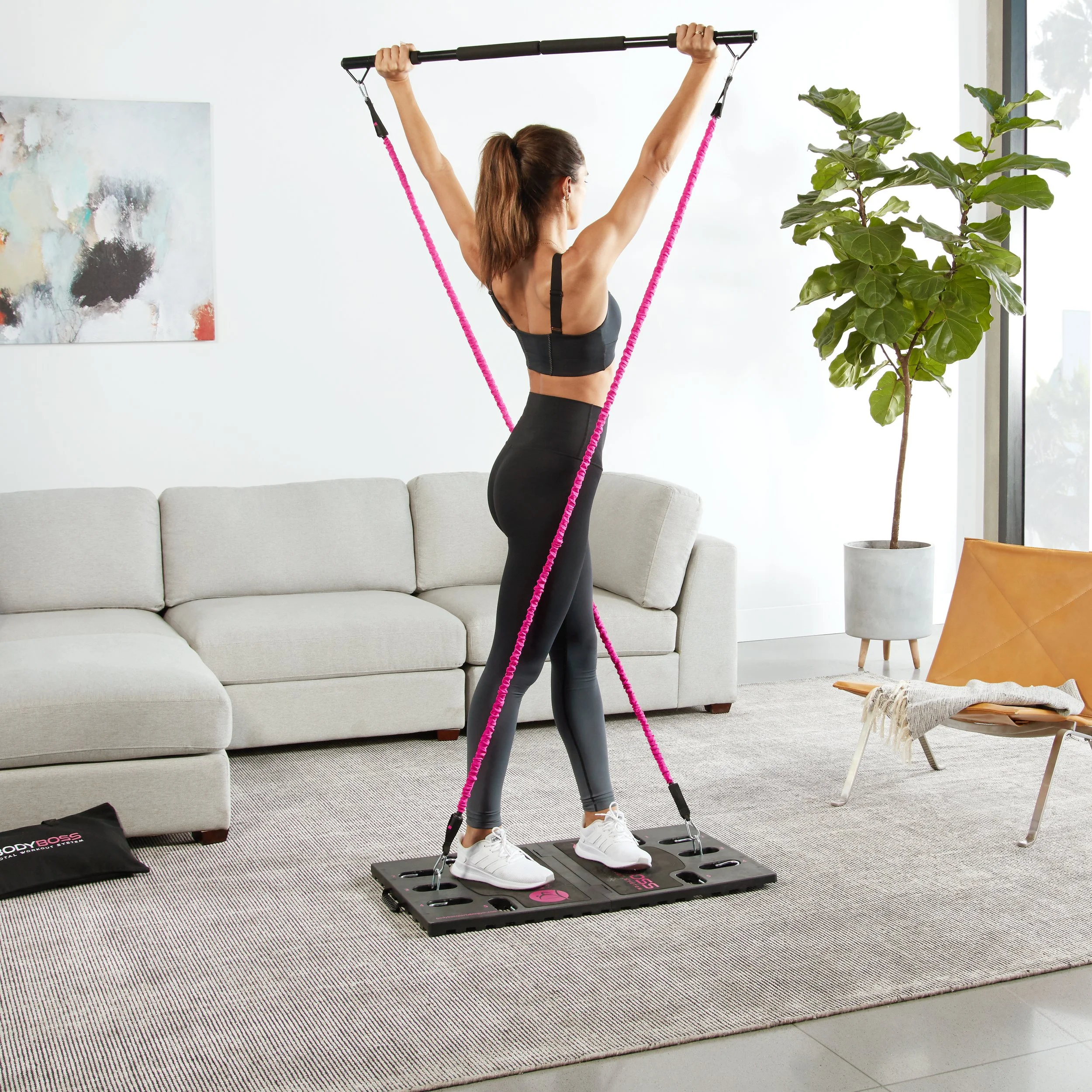 A woman in workout clothes stands on a rebounder, holding a stick above her head with resistance bands attached, in a living room with a gray sofa, a large potted plant, and abstract art on the wall.