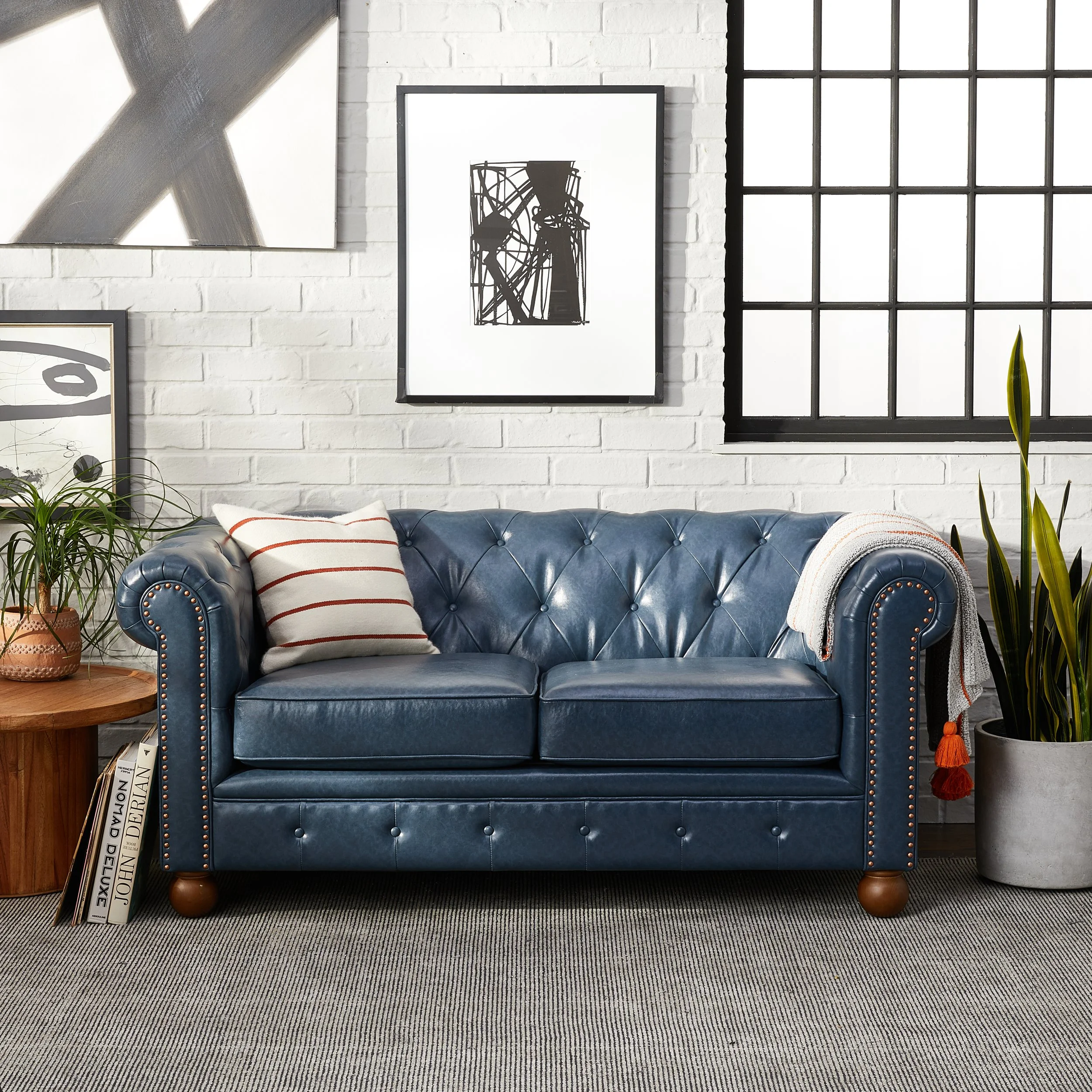 A stylish living room with a dark blue leather tufted sofa, a striped pillow, and a light textured blanket on the armrest. To the left, a small wooden side table holds a potted plant and books. The white brick wall behind features modern black-and-wh