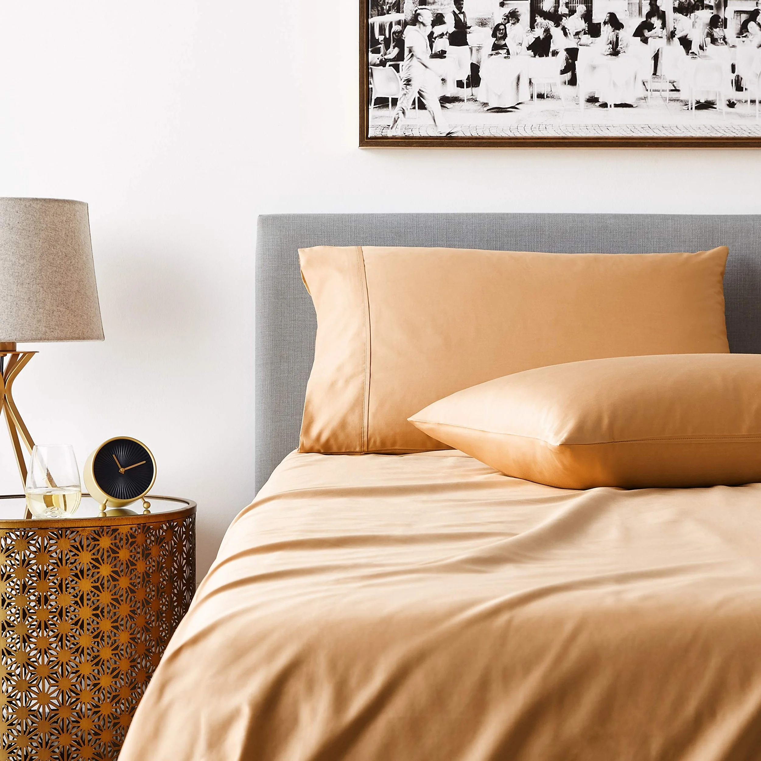 A neatly made bed with beige satin sheets and pillows, a grey upholstered headboard, a round wooden nightstand with a decorative cutout pattern, a glass of water, a black and gold clock, a beige lampshade, and a black and white framed photo on the wa
