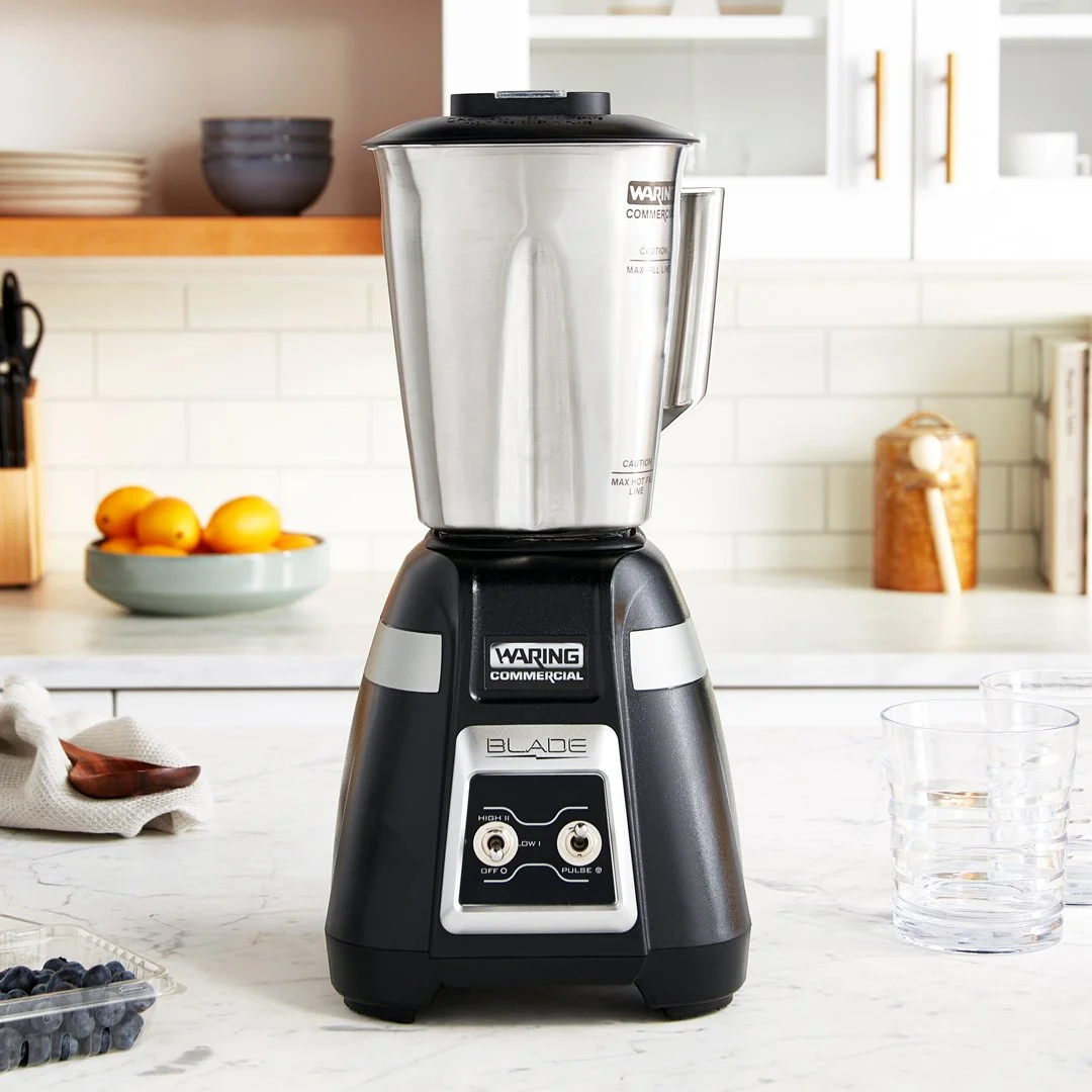 A commercial blender on a kitchen countertop with glasses of water, a bowl of blueberries, and a dish towel nearby. White cabinets and a bowl of lemons are in the background.