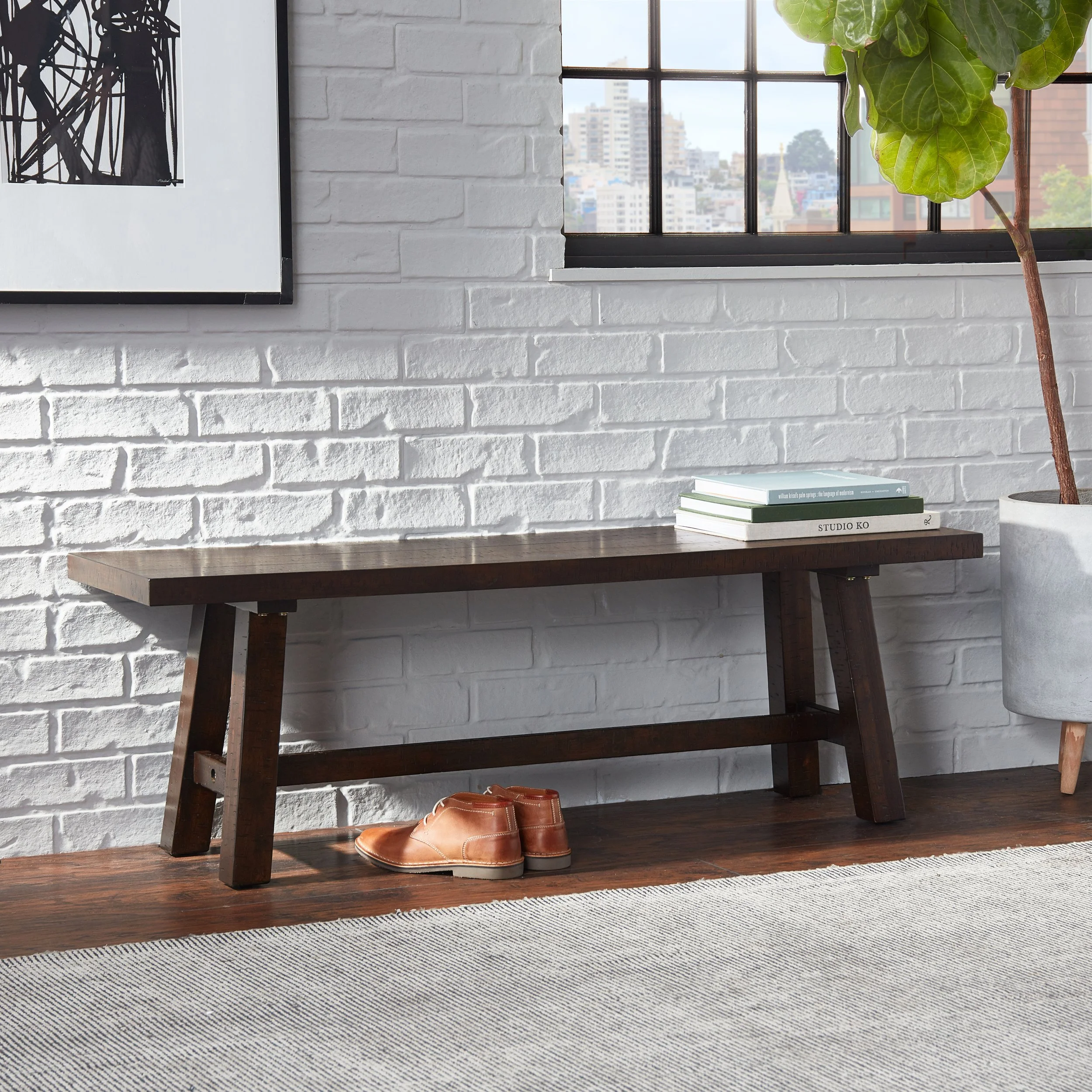 A dark wooden bench against a white brick wall, with a pair of brown leather shoes underneath, and a stack of books on the right side of the bench. A large plant in a white pot is on the far right, next to a window with black metal framing, overlooki