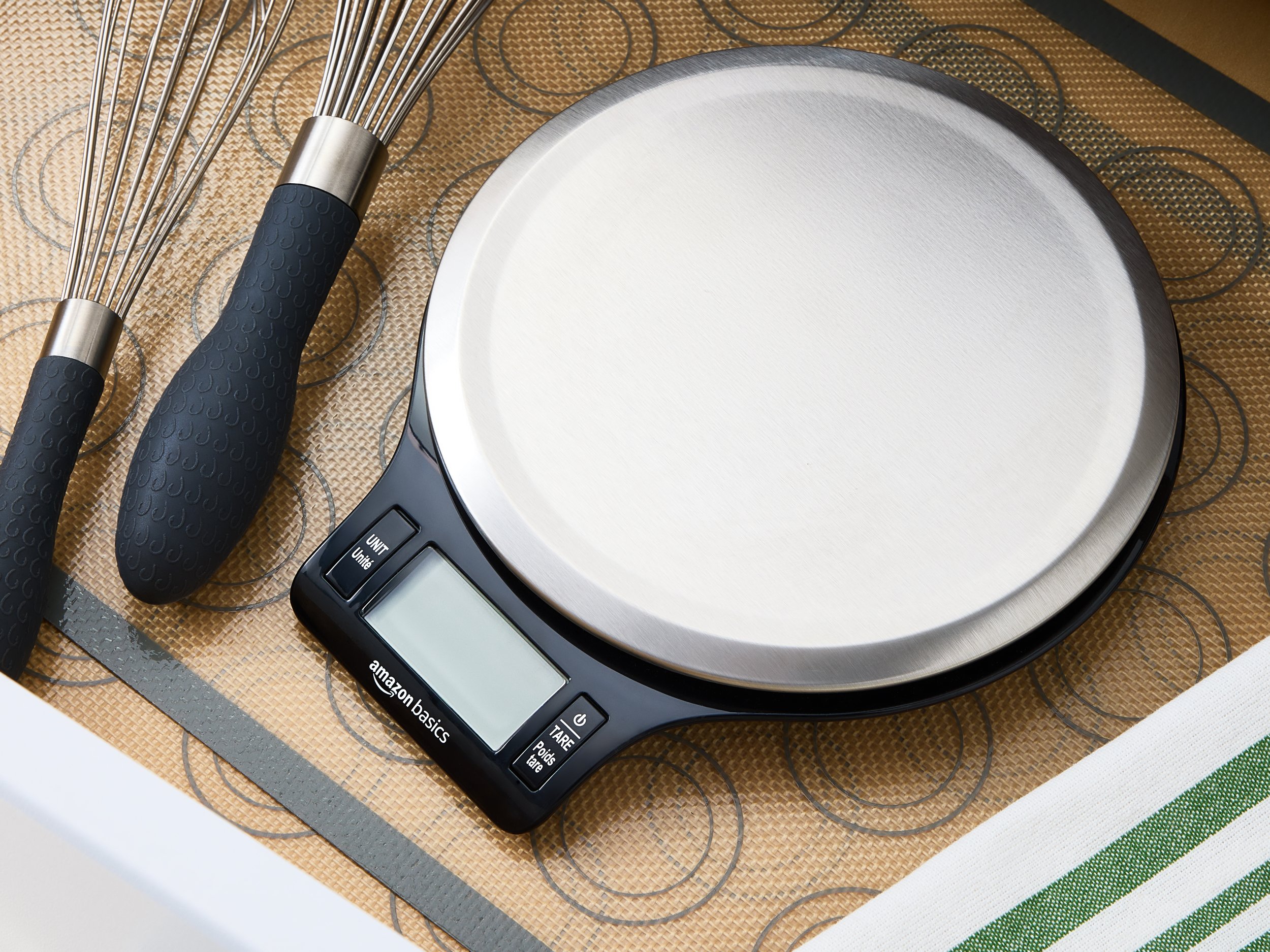 A digital kitchen scale sits on a countertop with a woven placemat underneath. Two whisks with black handles are placed beside the scale.