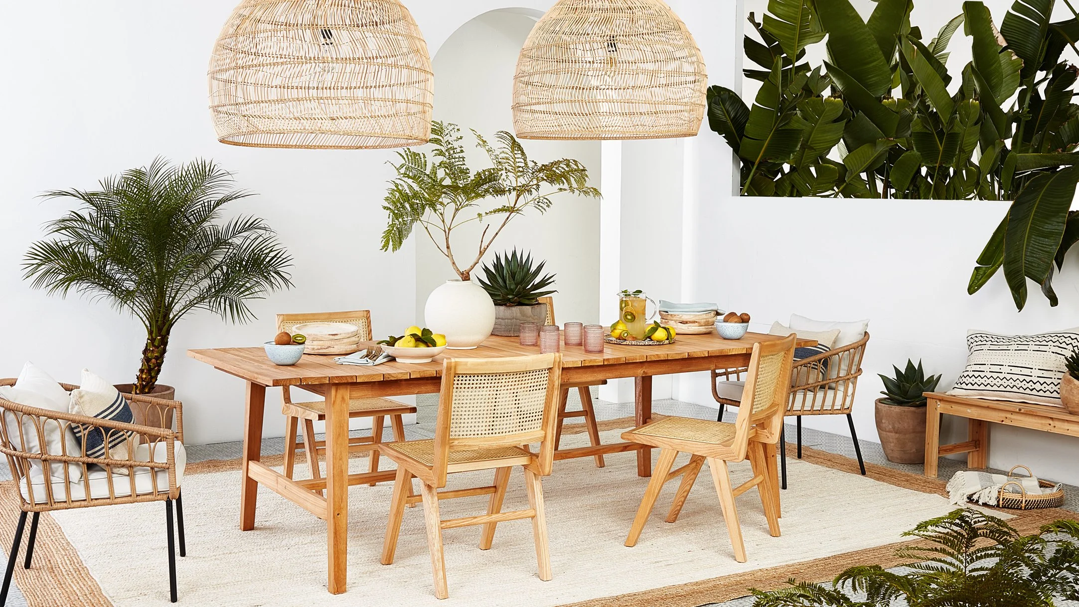 A bright dining room with a large wooden table surrounded by various chairs, decorated with large potted plants, hanging woven light fixtures, and a neutral-colored rug.