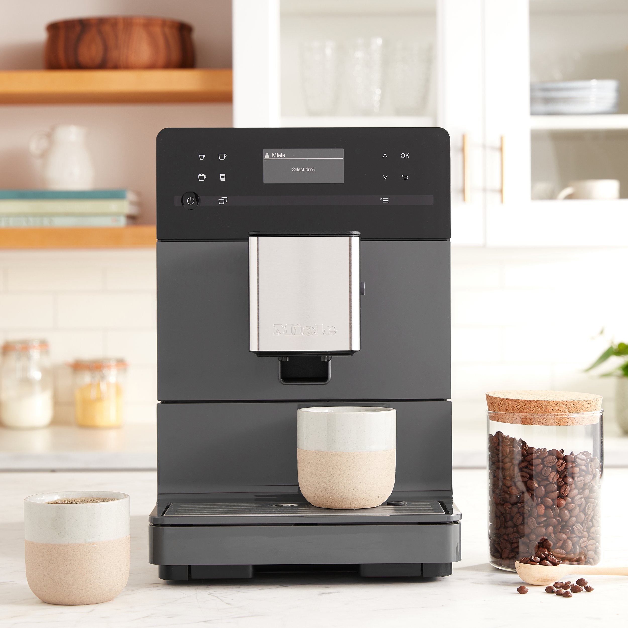 Modern coffee maker on a white kitchen counter, with coffee beans in a jar and two cups of coffee.