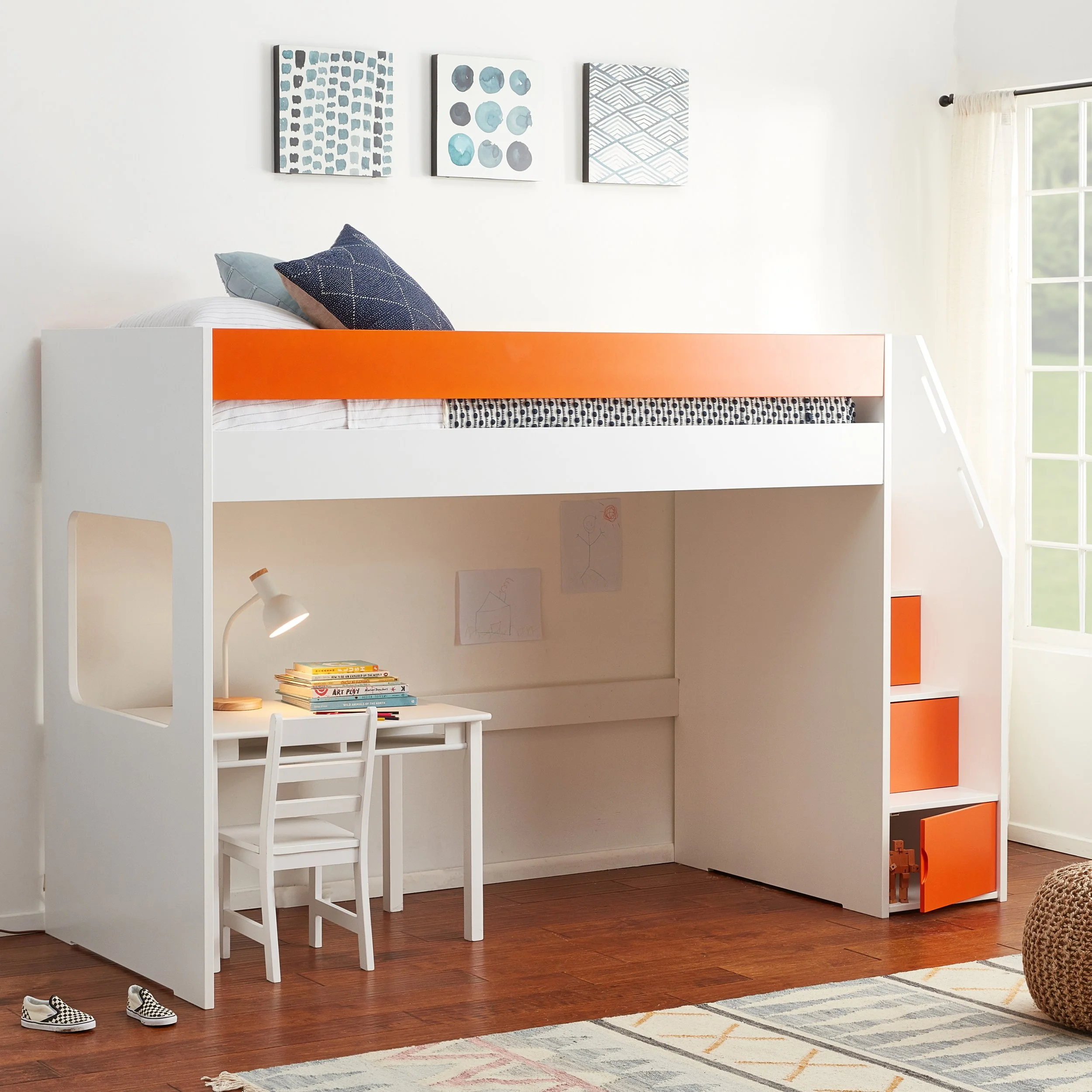 Child's bedroom featuring a loft bed with white and orange frame, a small white desk and chair underneath, a table lamp, books, and children's drawings on the wall, with a window and a woven basket nearby.
