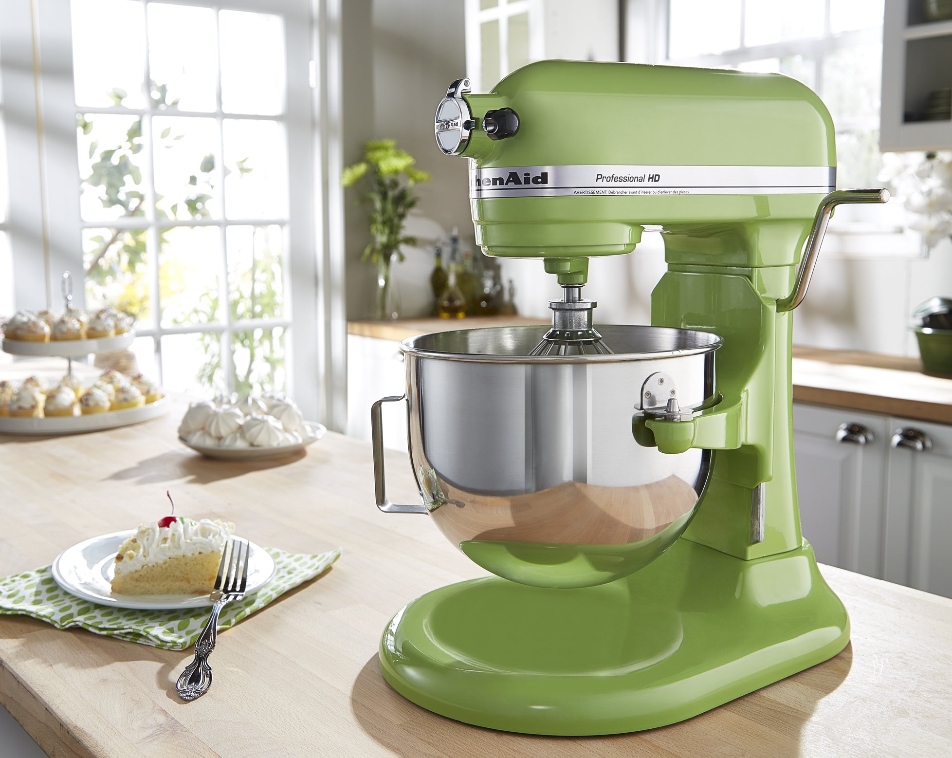 A bright green KitchenAid stand mixer on a wooden kitchen island, with a dessert plate containing a slice of cake, a fork, and a green napkin. In the background, there are cupcakes on a tiered serving tray and a window with sunlight streaming in.