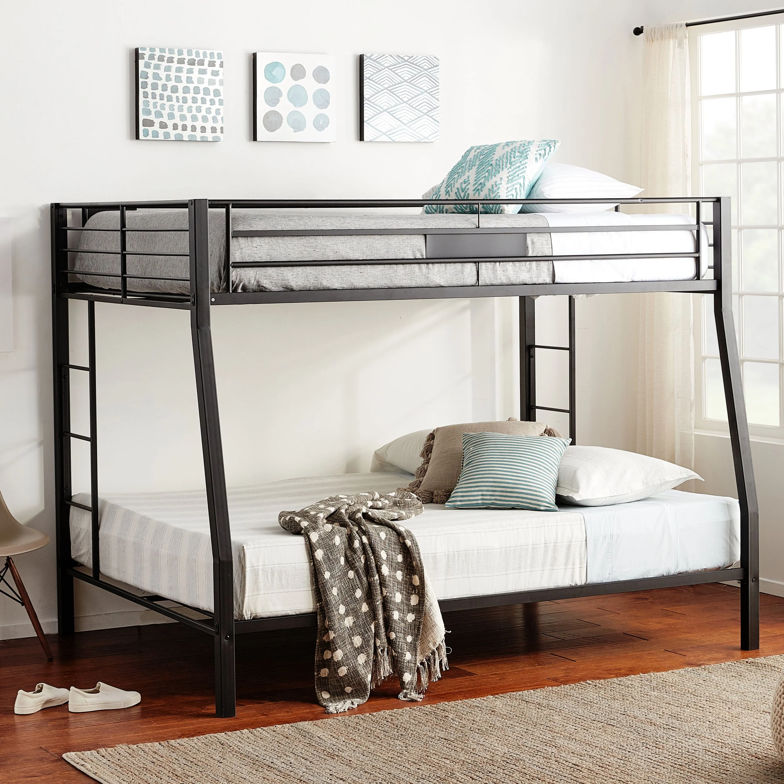 A bunk bed with a black metal frame in a bright bedroom, with white walls, a window with curtains, and three abstract art pieces on the wall. The lower bed has pillows and a blanket, while the upper bed has pillows and a mattress.
