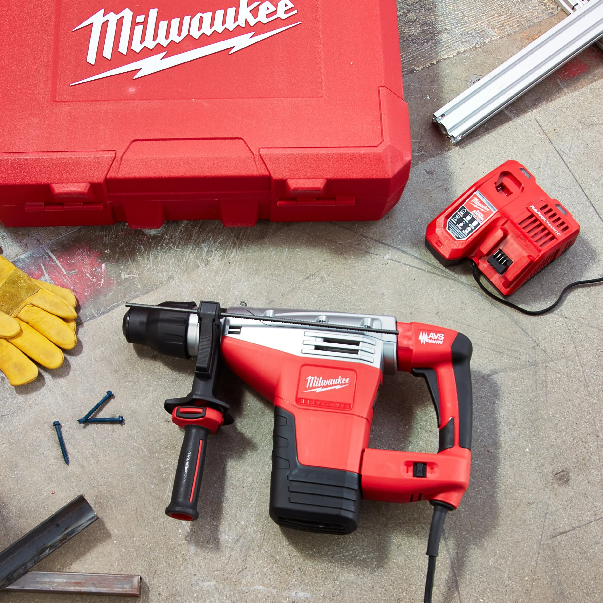 A Milwaukee rotary hammer drill, a red Milwaukee toolbox, a red Milwaukee battery charger, some yellow work gloves, loose blue screws, and a metal pipe on a concrete floor.