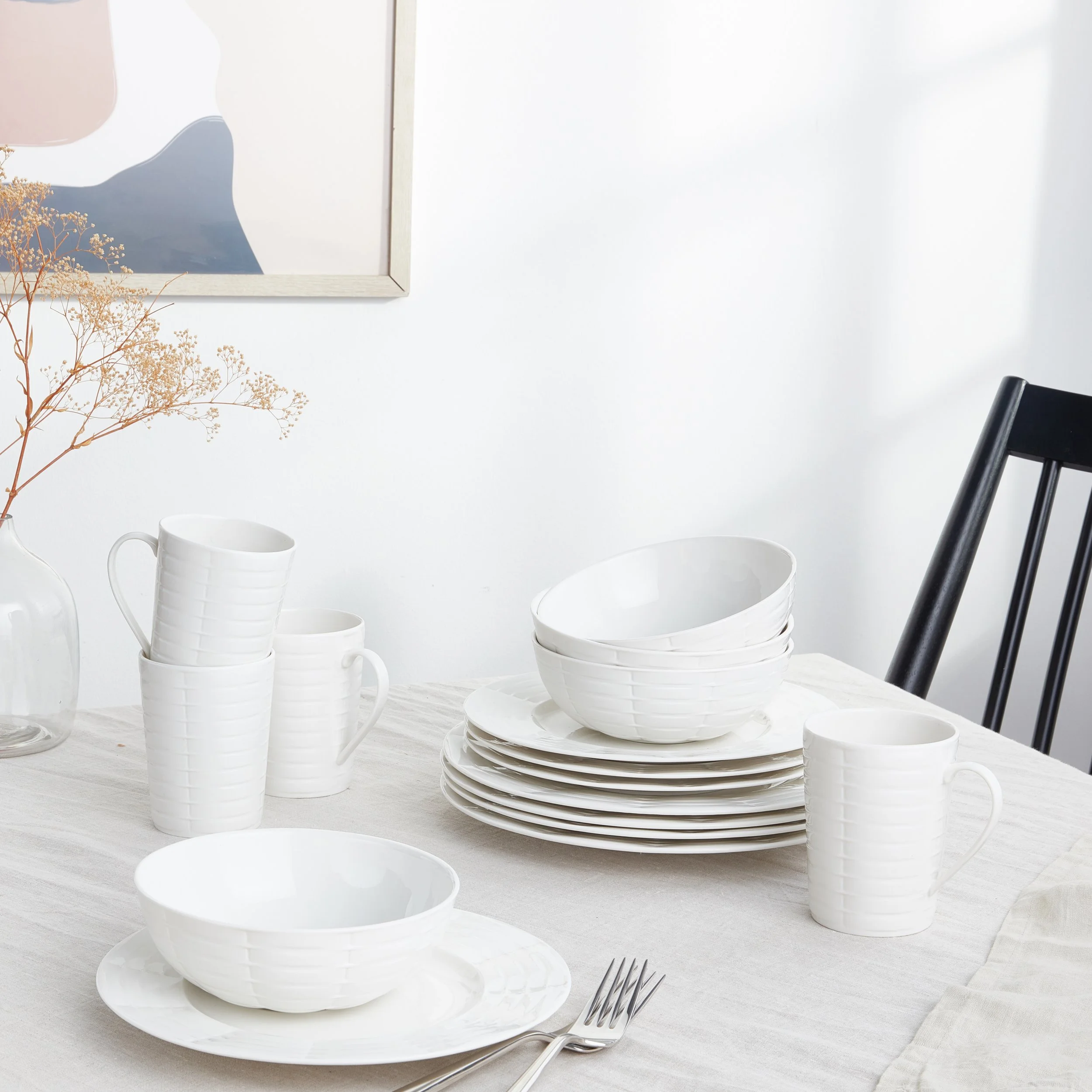 Stacked white ceramic bowls and cups on a dining table with a fork, a flower in a vase, and a black chair in the background.