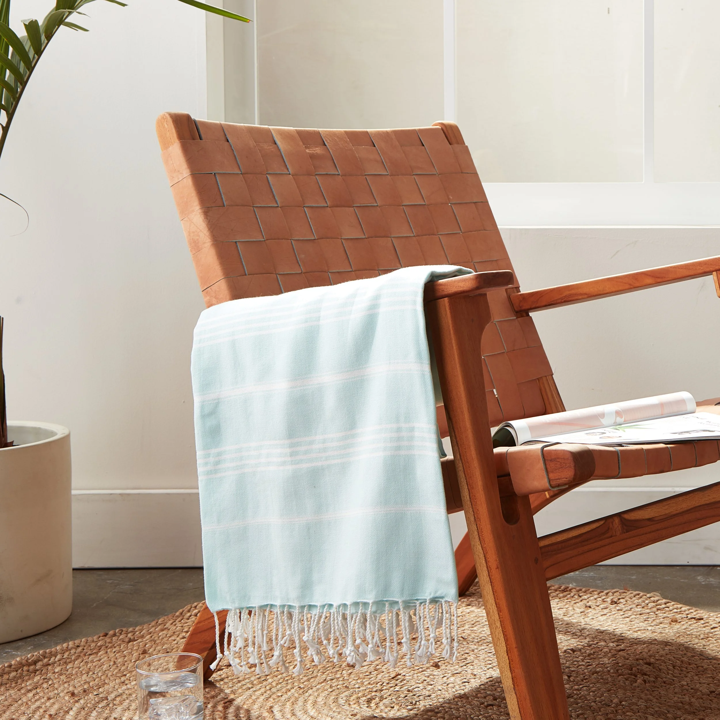A wooden lounge chair with a brown woven leather seat and backrest, draped with a light blue striped towel. There is a glass of water on the floor next to the chair, and a potted plant partially visible on the left side. The setting appears to be nea