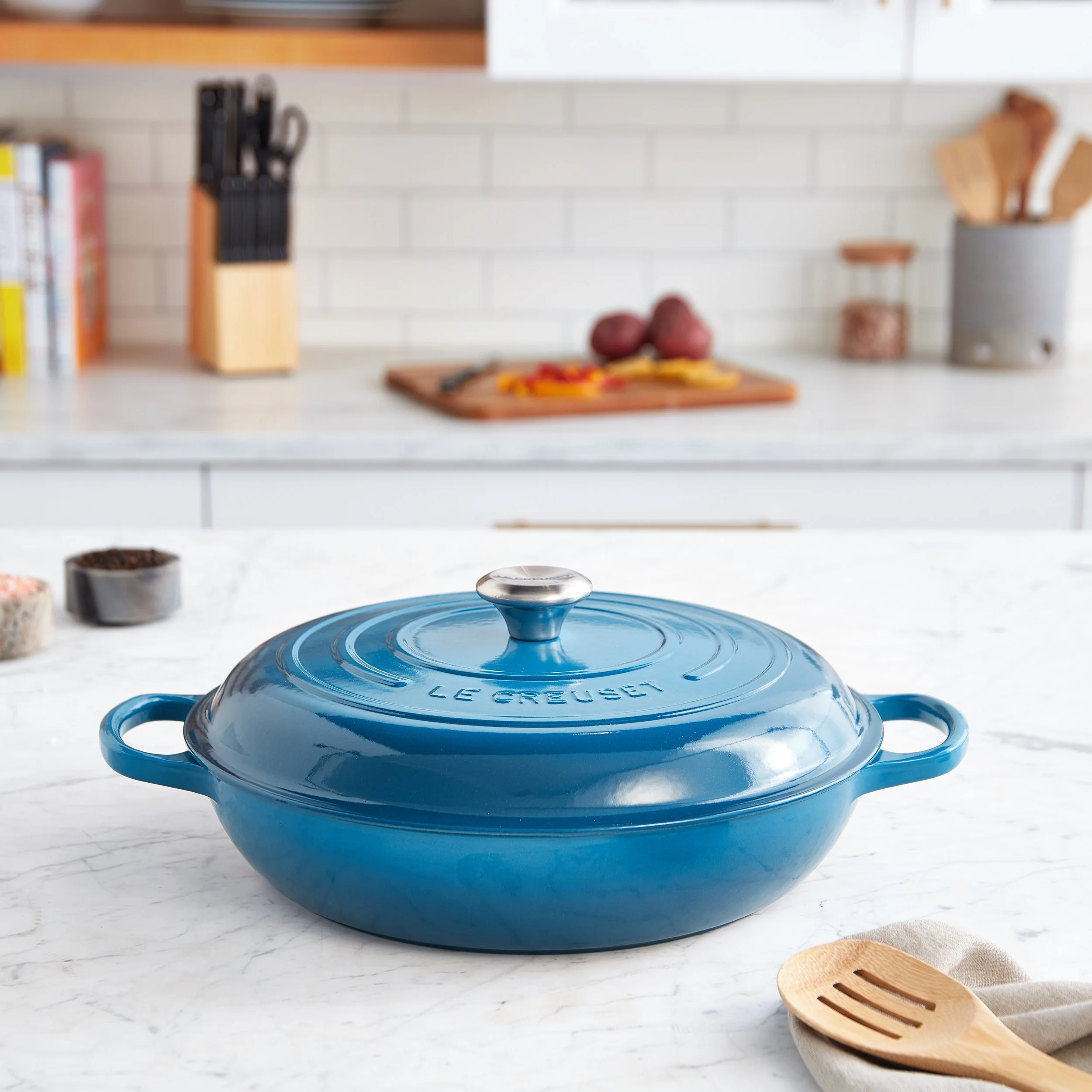 A blue Le Creuset cast iron casserole dish with a lid on a white marble countertop. In the background, kitchen items including a knife block, cutting board with vegetables, spices, and a utensil holder are visible.