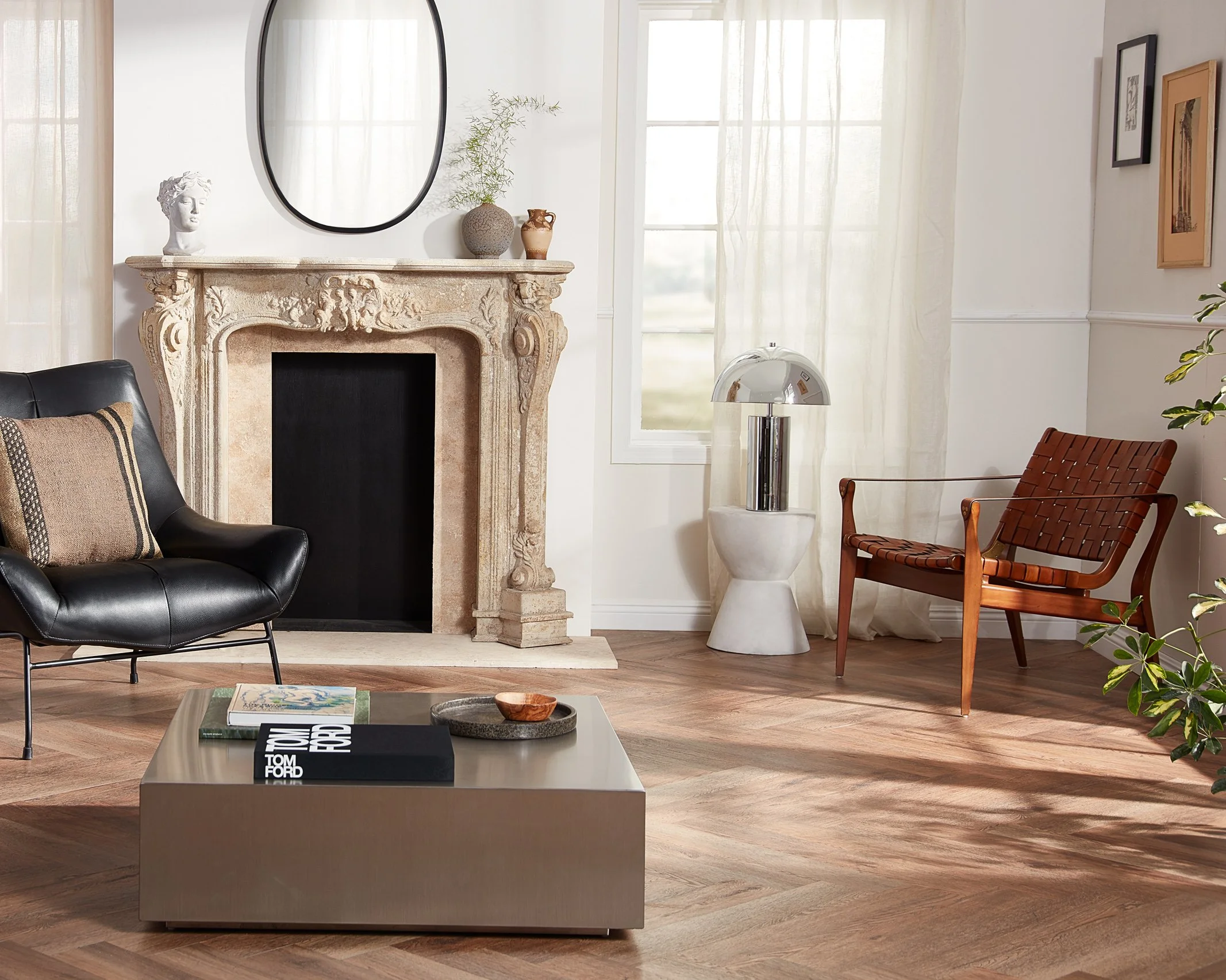 Living room with a stone fireplace, black leather armchair with a pillow, wooden lounge chair, rectangular coffee table with books and decorative items, a contemporary white table lamp, and framed artwork on the walls, filled with natural light from 