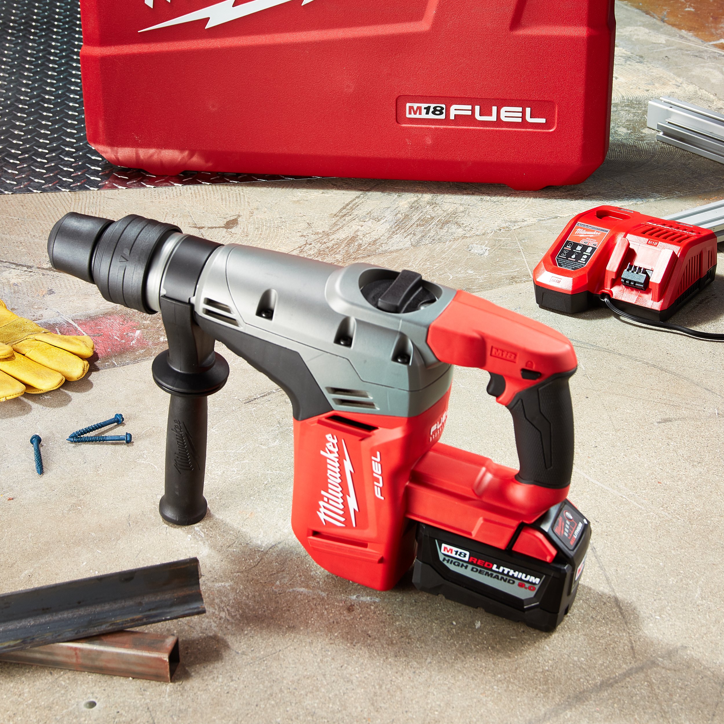 A Milwaukee FUEL Cordless Rotary Hammer with a red and black battery pack on a workbench. In the background, there is a red Milwaukee M18 FUEL box and an M18 battery charger. Yellow work gloves and some scattered screws are also visible on the work surface.