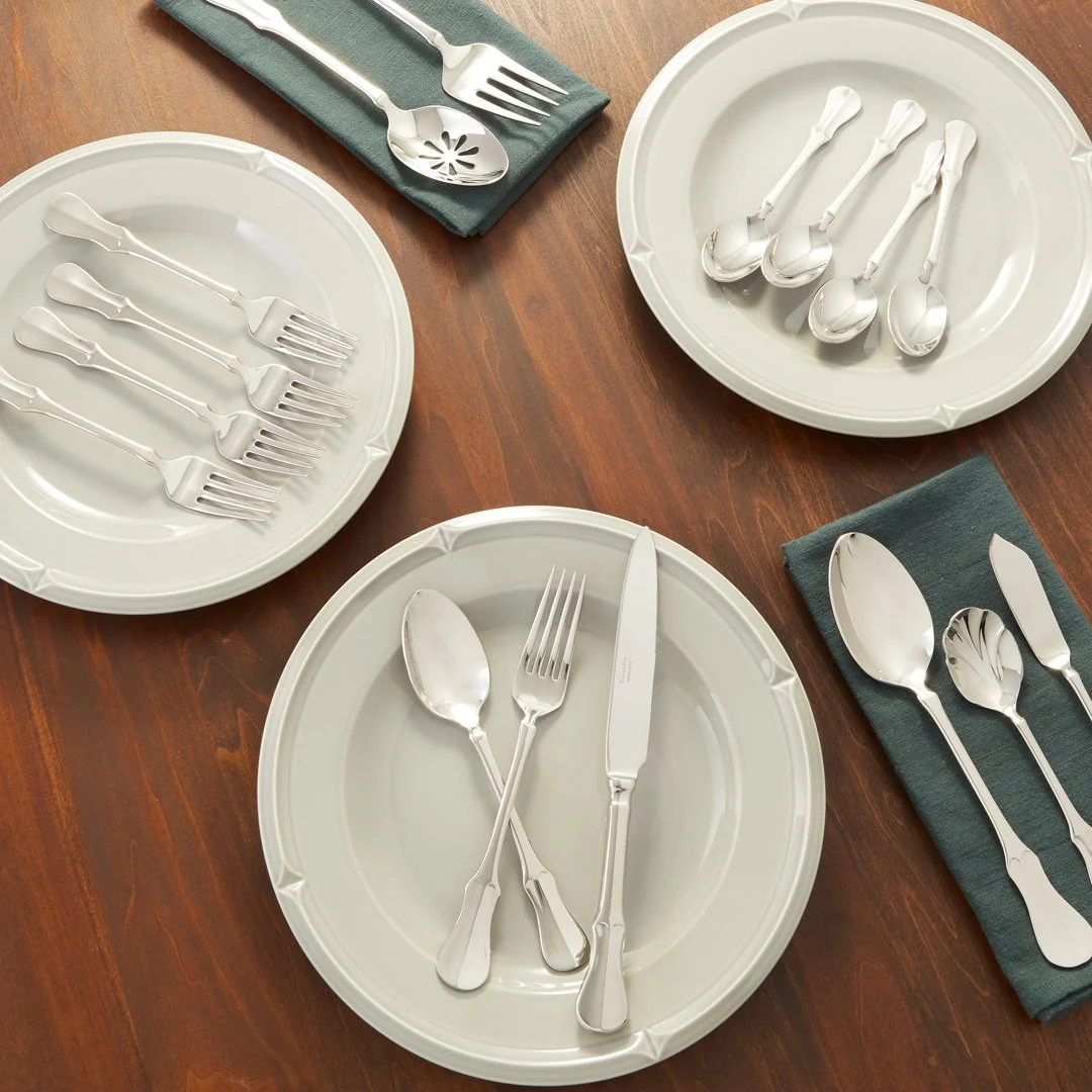 Tablesetting with white plates, silver utensils including forks, knives, and spoons, and teal napkins on a wooden table.