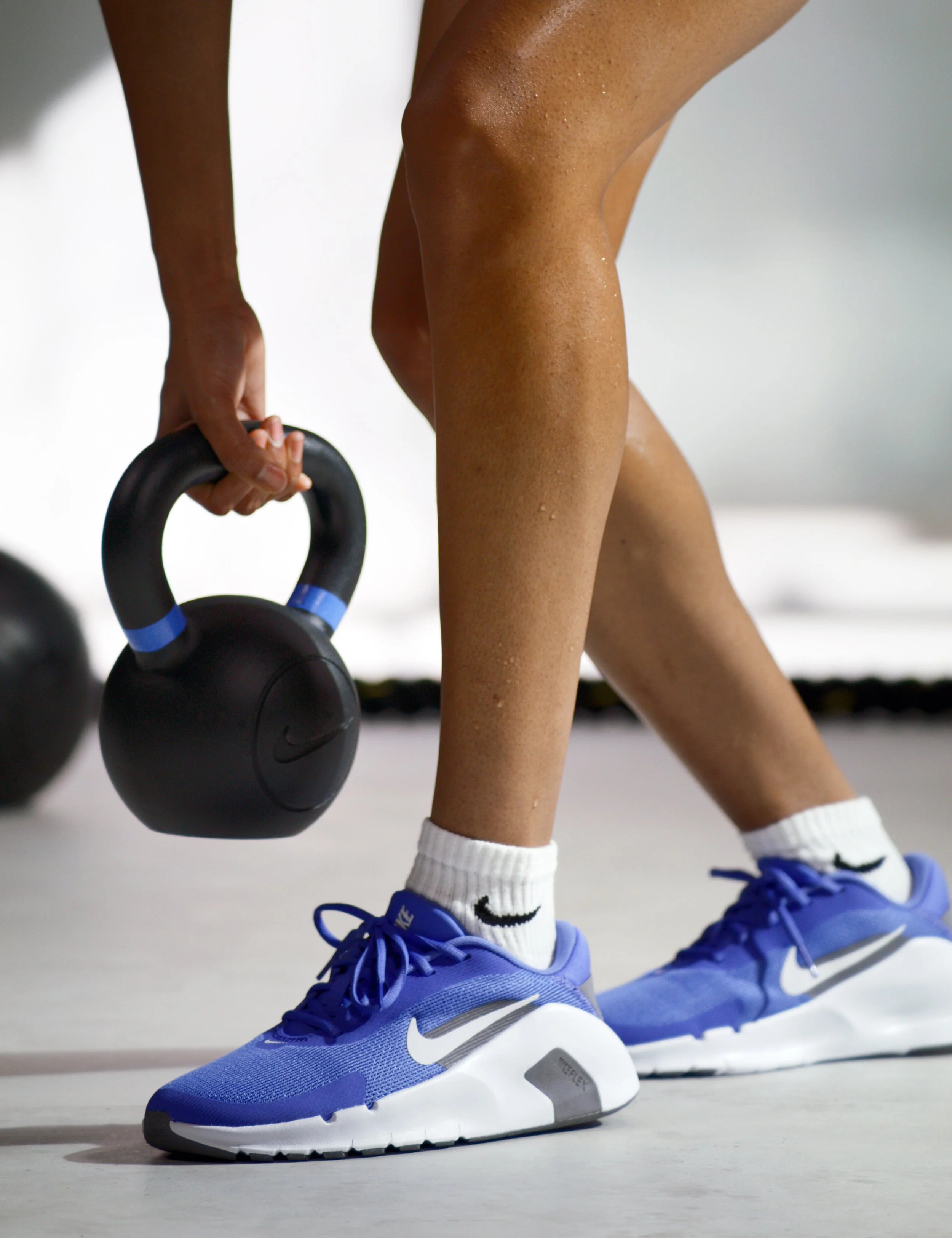 Person in blue Nike sneakers and white Nike socks with black swoosh, holding a black kettlebell in a gym.