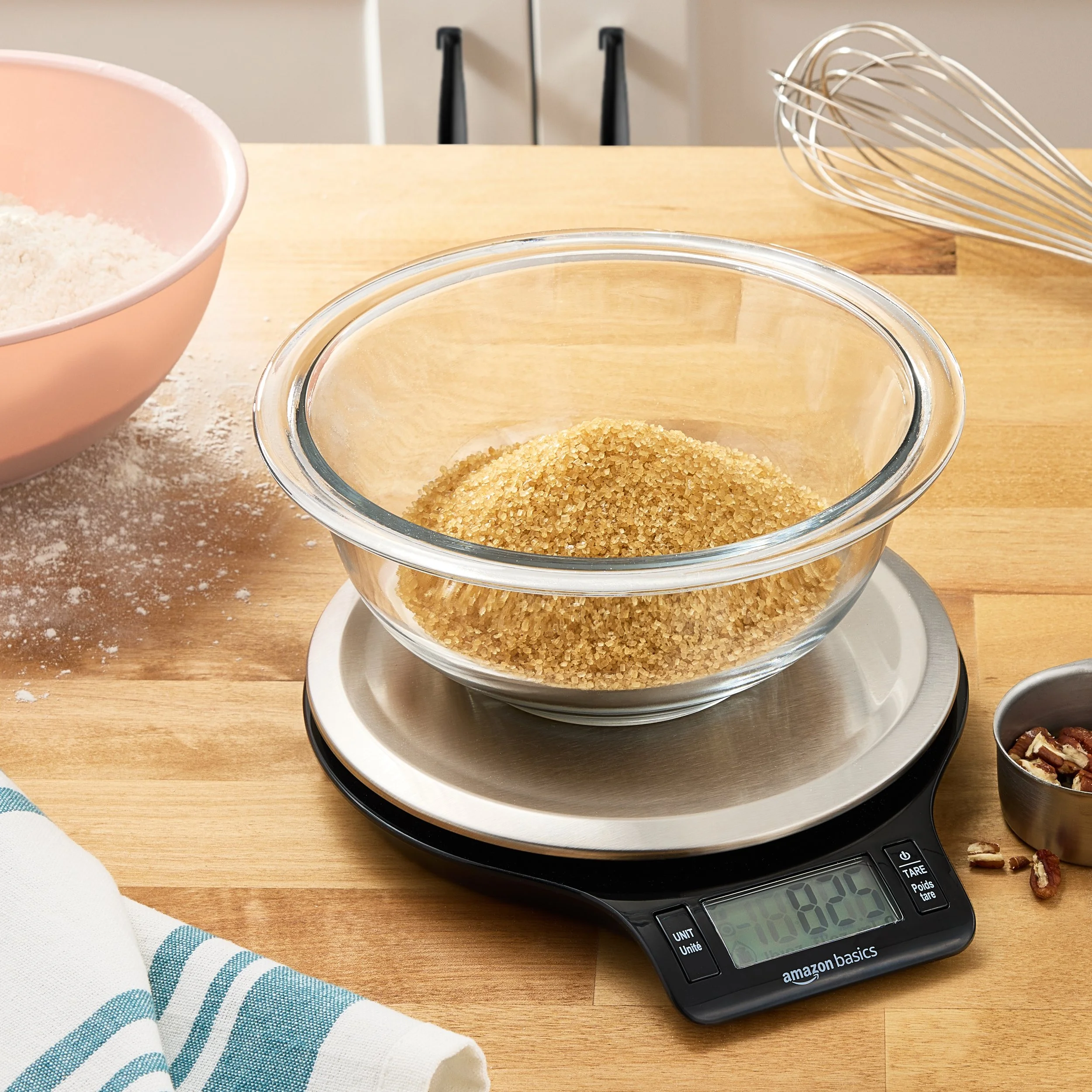 A glass mixing bowl of brown sugar on a kitchen scale, with baking ingredients and utensils on a wooden countertop.