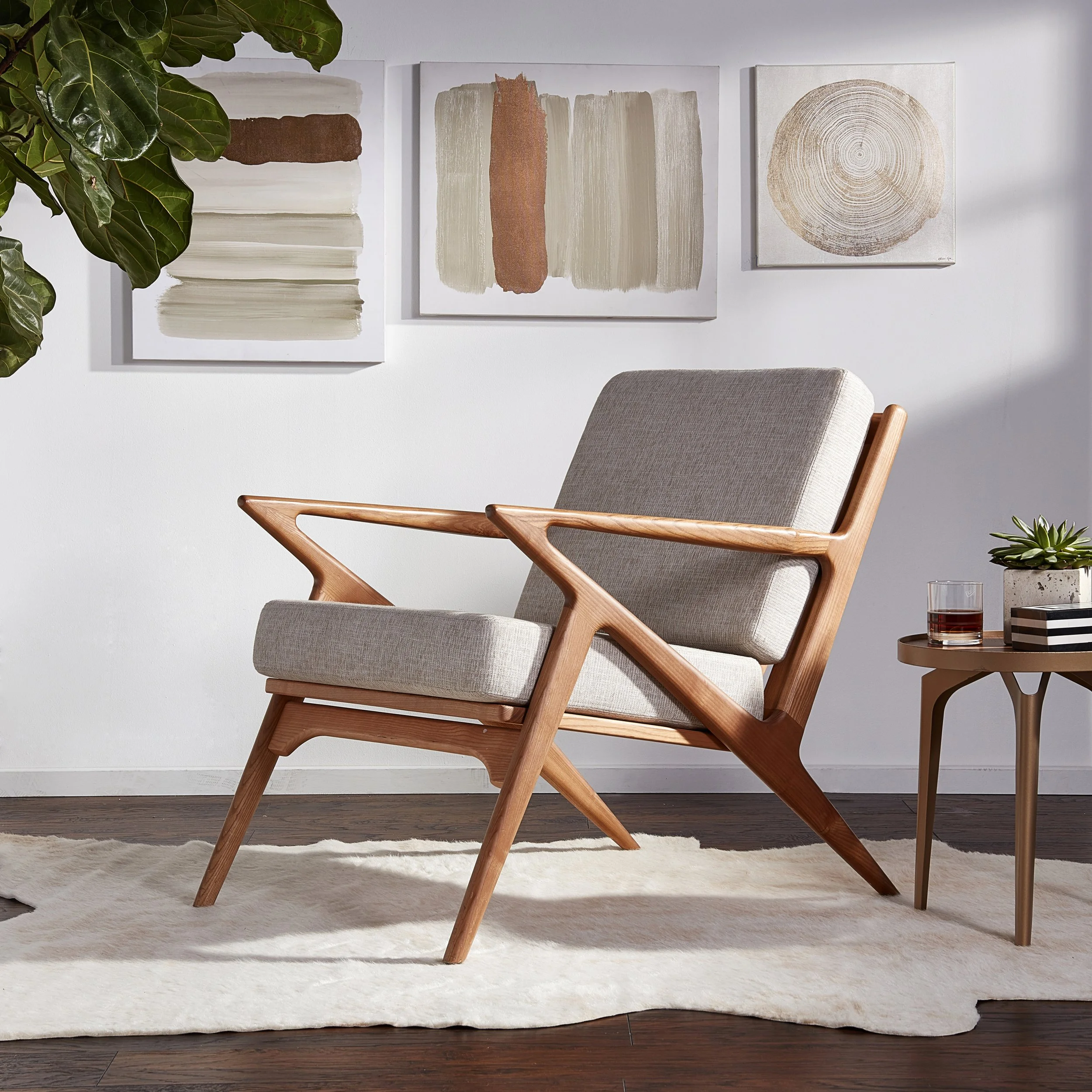 Modern living room with beige armchair featuring wooden frame, white wall with abstract art, a side table with a glass of drink and a potted plant, and a cream-colored rug on dark wooden flooring.