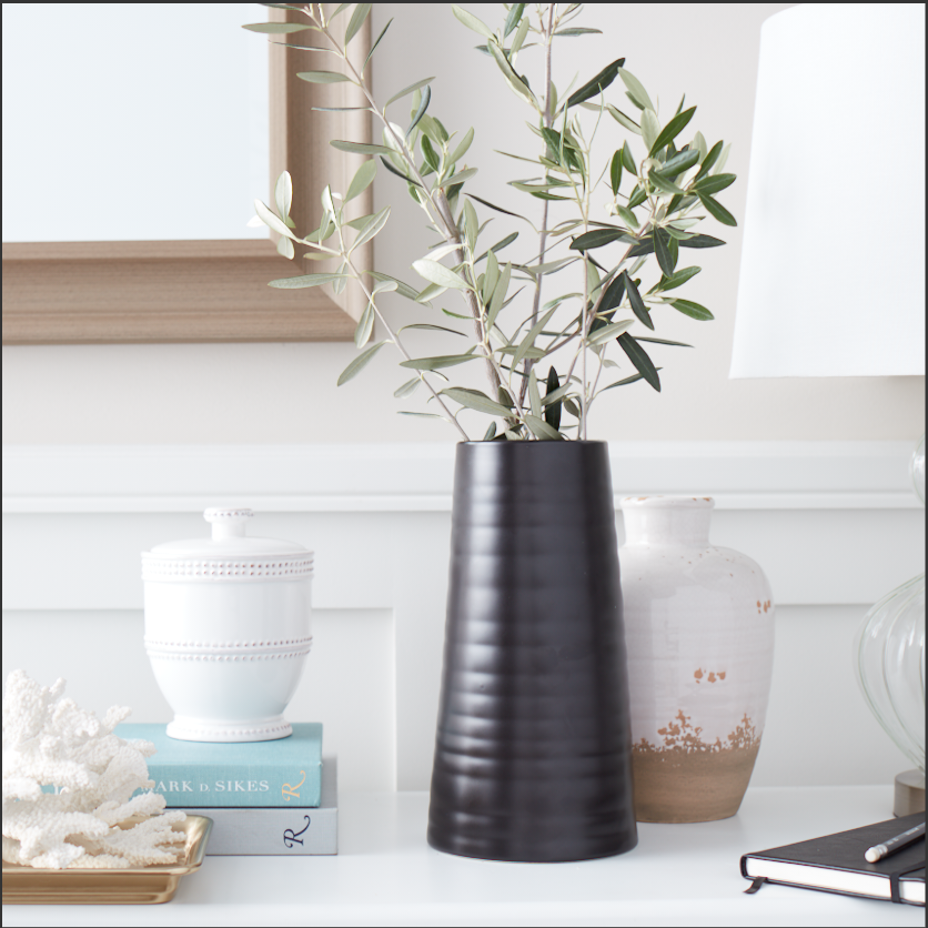 A black ribbed vase with green leafy branches on a white surface, flanked by a white ceramic jar with a lid, a beige and white decorative vase, and a small stack of books, with a mirror and white wall in the background.