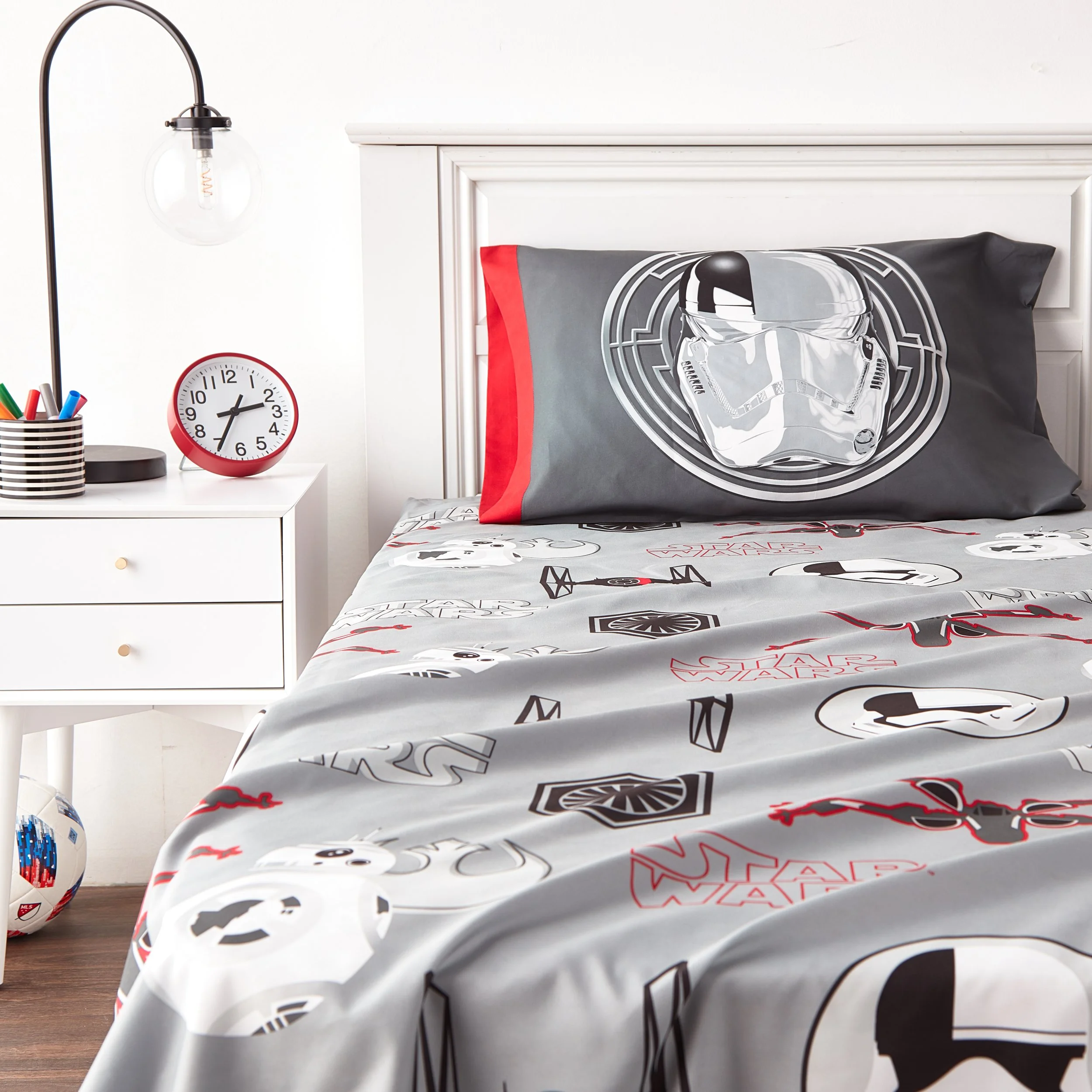 Star Wars themed bedroom with a bed, pillow, and blanket featuring Star Wars characters and symbols, a white nightstand with pens and a clock, a wall-mounted lamp, and a soccer ball on the floor.