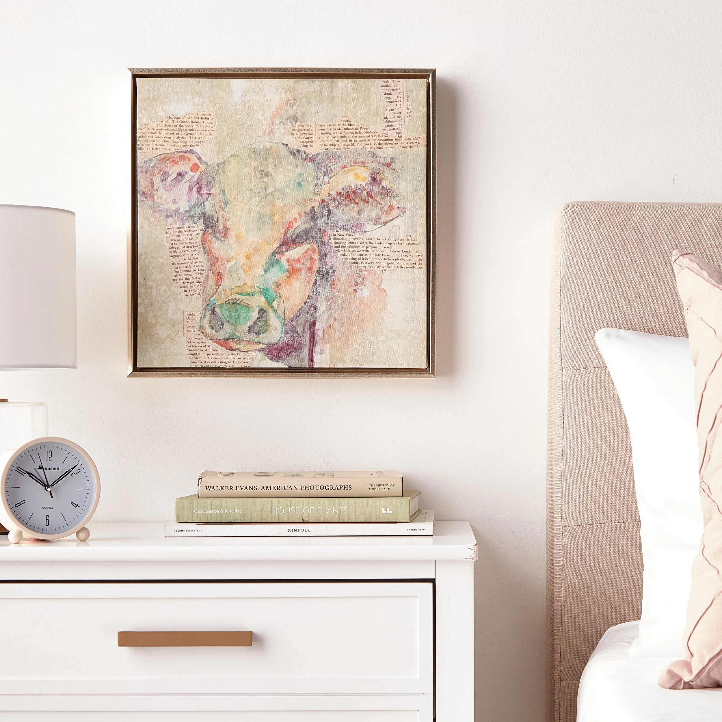 A bedroom nightstand with a stack of books, a clock showing 9:10, and a lamp. Above the nightstand is a framed watercolor painting of a cow on a textured background.