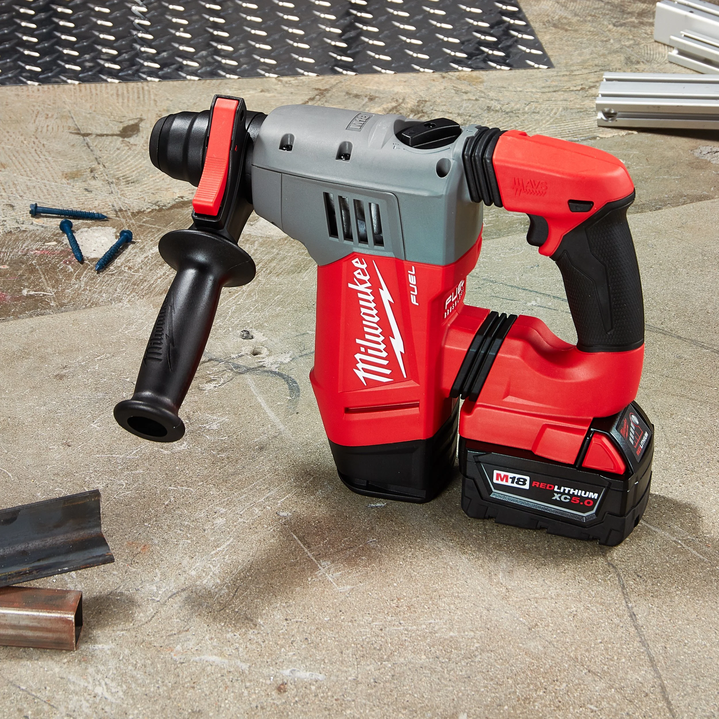 A Milwaukee cordless rotary hammer drill with a red and black lithium-ion battery attached is on a work surface at a construction site, with some screws, metal pieces, and a rubber mat in the background.