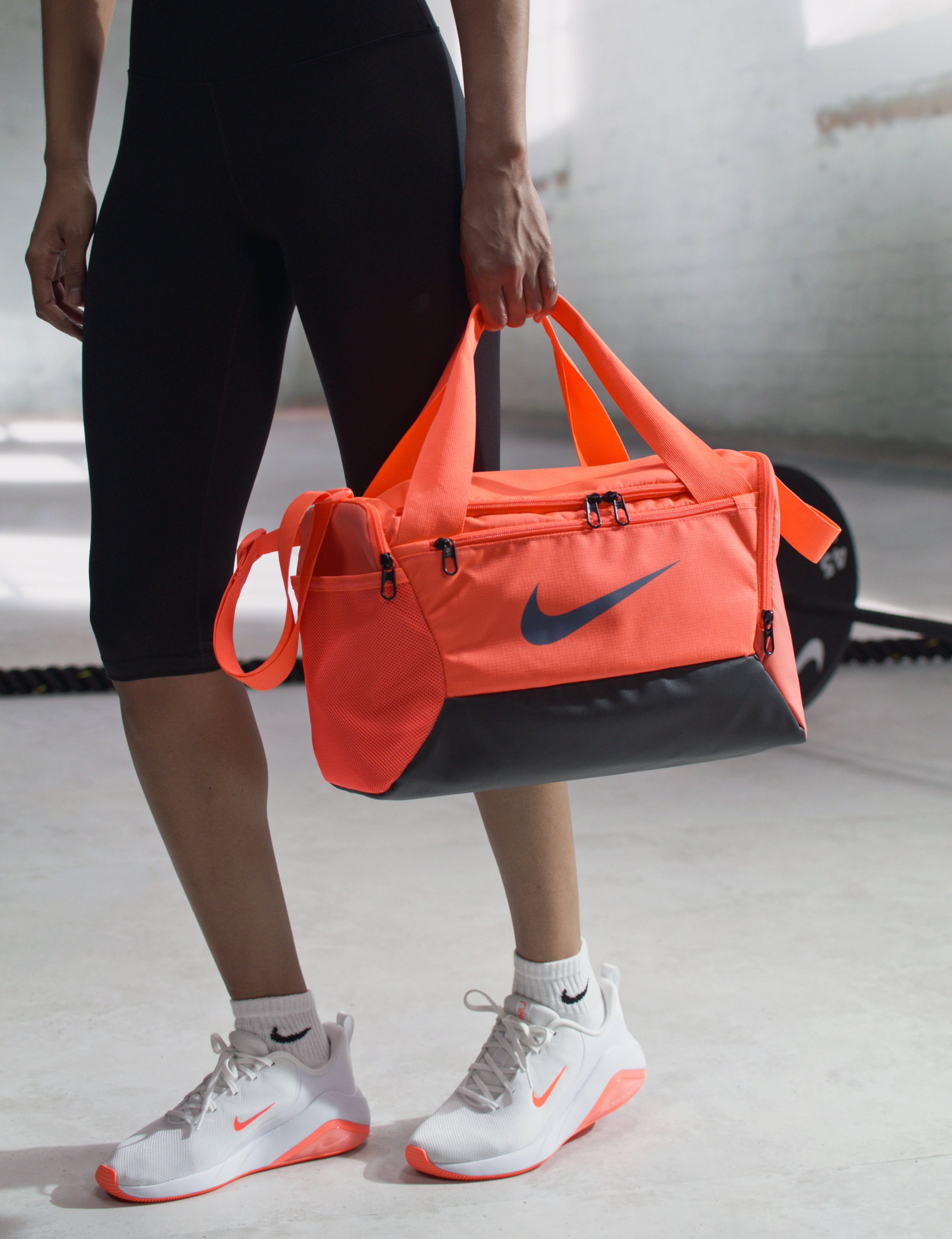 A person wearing white athletic shoes and black athletic pants, holding a bright orange Nike gym bag with black accents inside a gym.