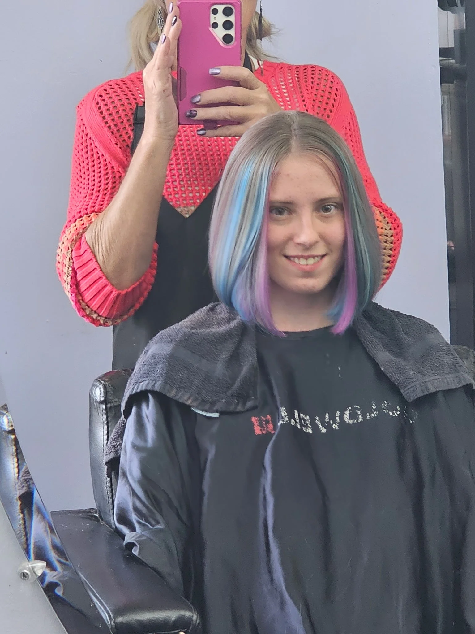 Young woman with guitar-shaped rainbow-colored hair getting a haircut at a salon, sitting in a chair with a black cape, while her stylist takes a selfie in the mirror using a pink phone. Radiant Styles by Julie
