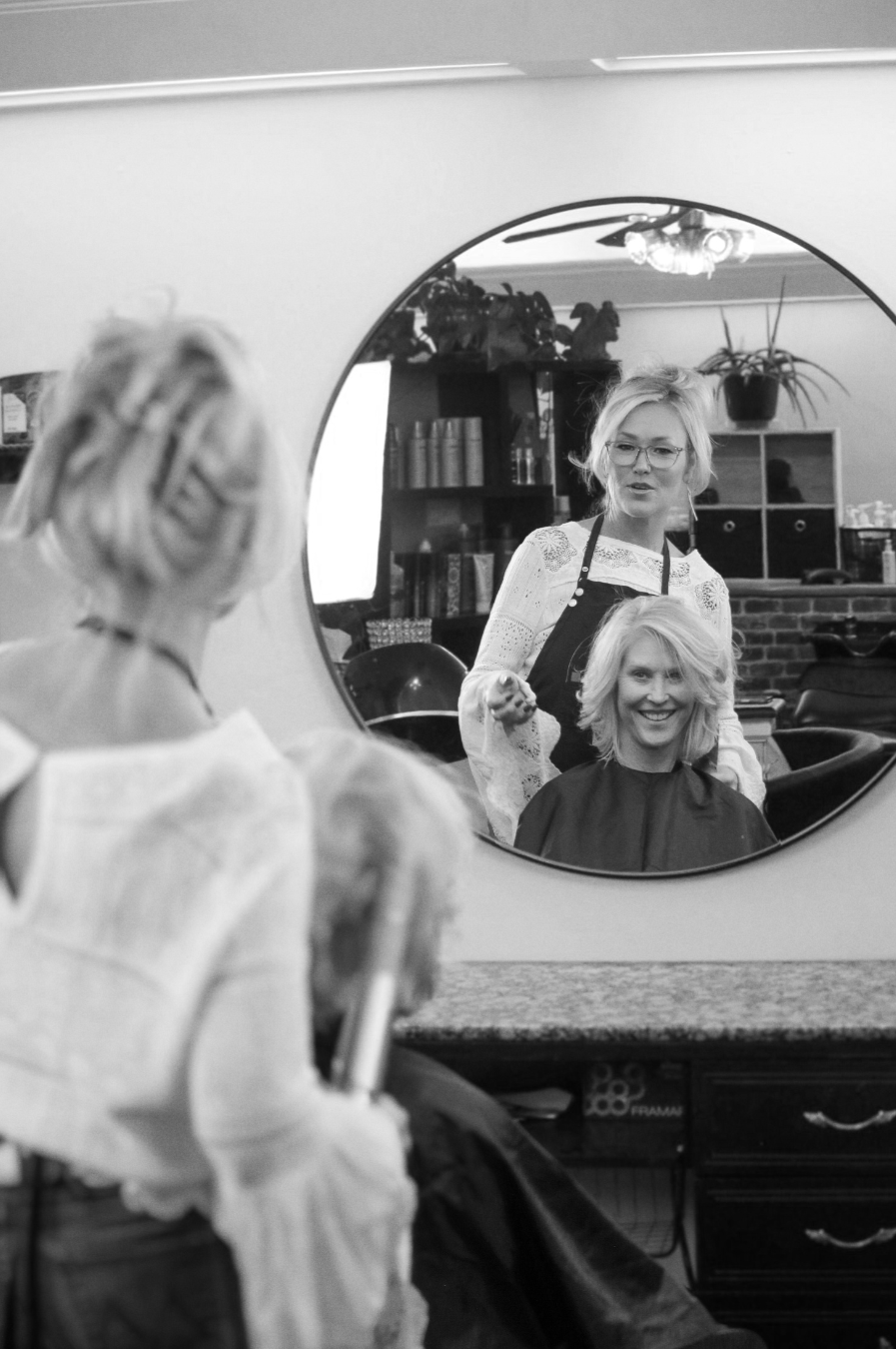 A woman getting her hair styled at a salon, looking at her reflection in a large round mirror, with the hairstylist standing behind her smiling. Julie, the founder of Radiant Styles by Julie