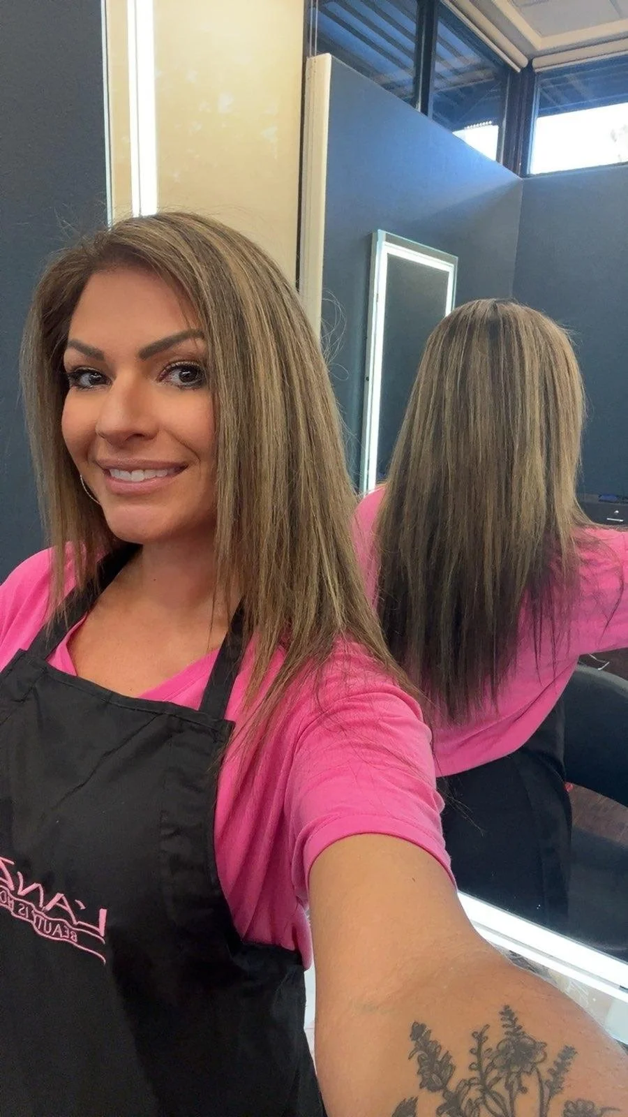 Woman with shoulder-length light brown hair taking a selfie in a salon, wearing a pink shirt and a black apron, with a tattoo of flowers on her forearm, and a mirror reflecting her back. Radiant Styles by Julie
