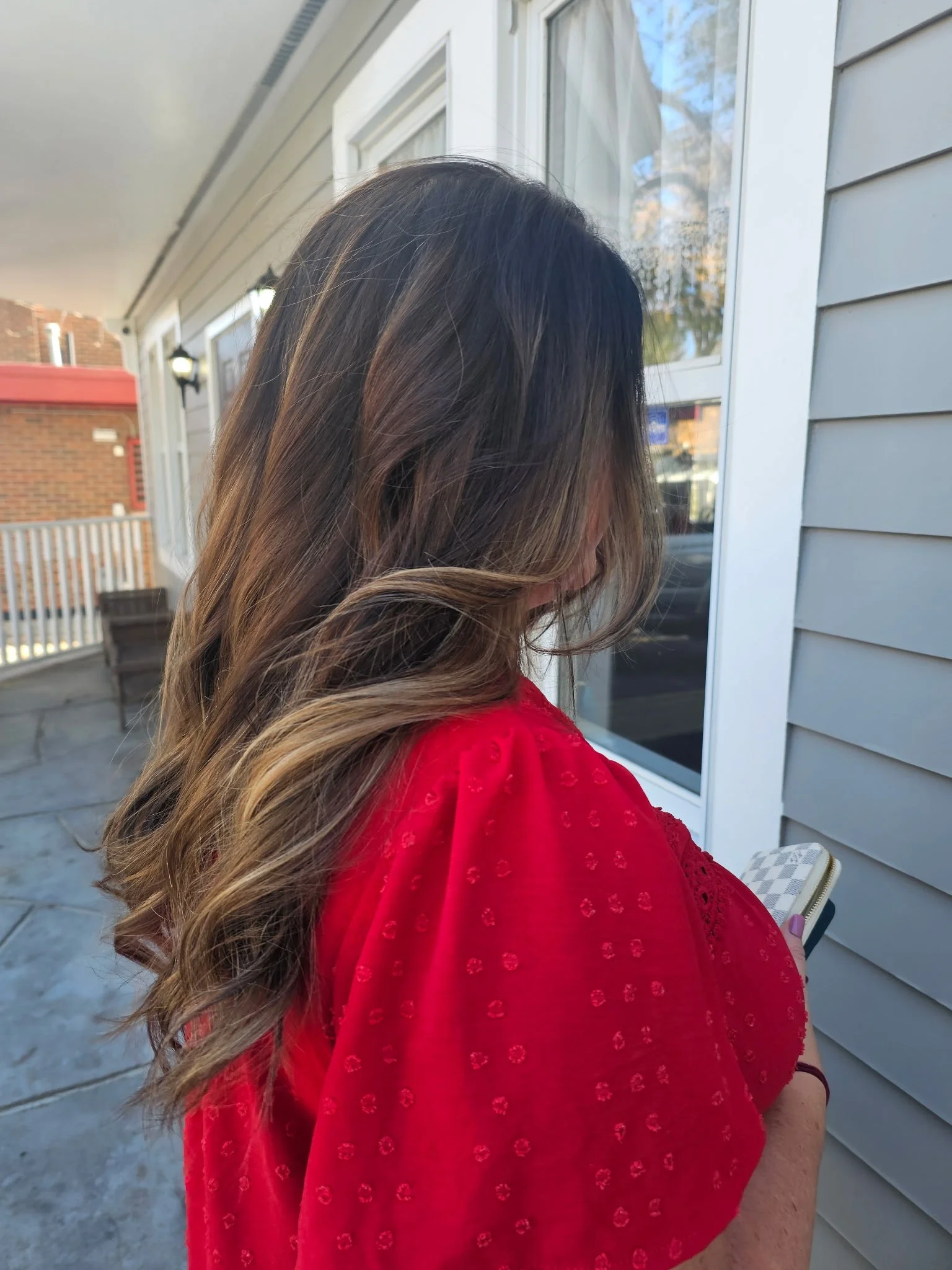 Woman with long brown hair wearing a red blouse standing on a patio near a house with light gray siding, just had her hair done by Radiant Styles by Julie