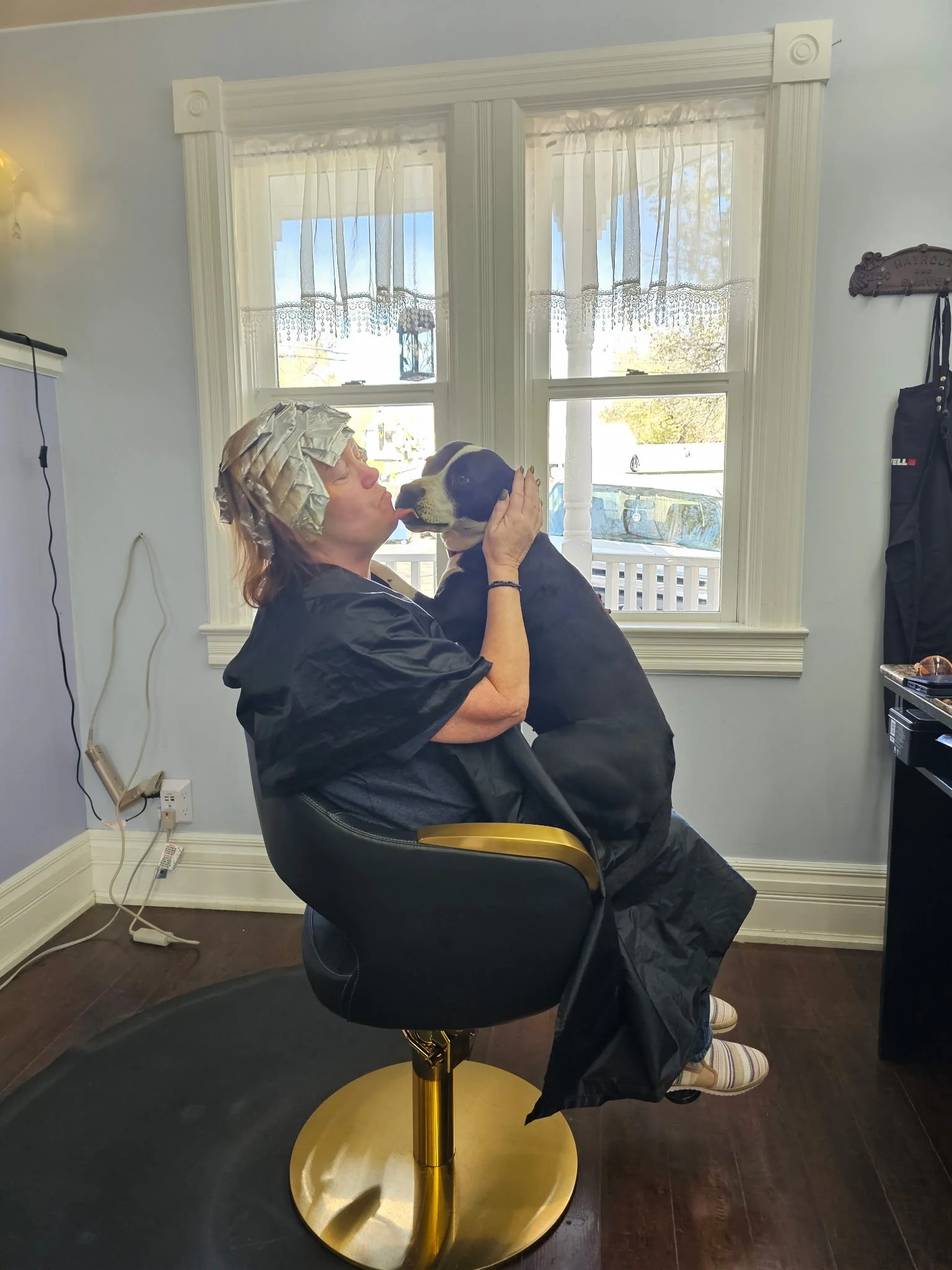 A woman sitting in a barber chair with a towel wrapped around her hair in a color, wearing a black cape, is giving a kiss to a black and white dog in front of her, inside a room with a window. Radiant Styles by Julie