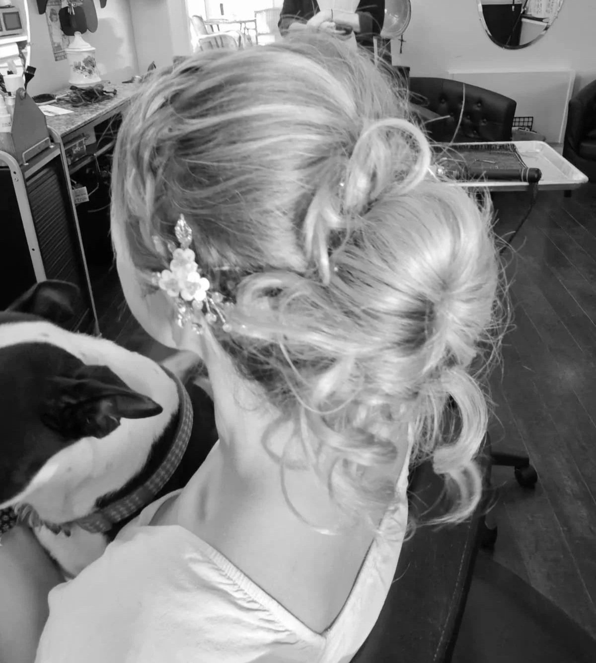 A woman with elegantly styled blonde hair adorned with a floral hair accessory, sitting in a salon chair, holding a dog with a collar. The salon has various supplies and a mirror in the background. Radiant Styles by Julie
