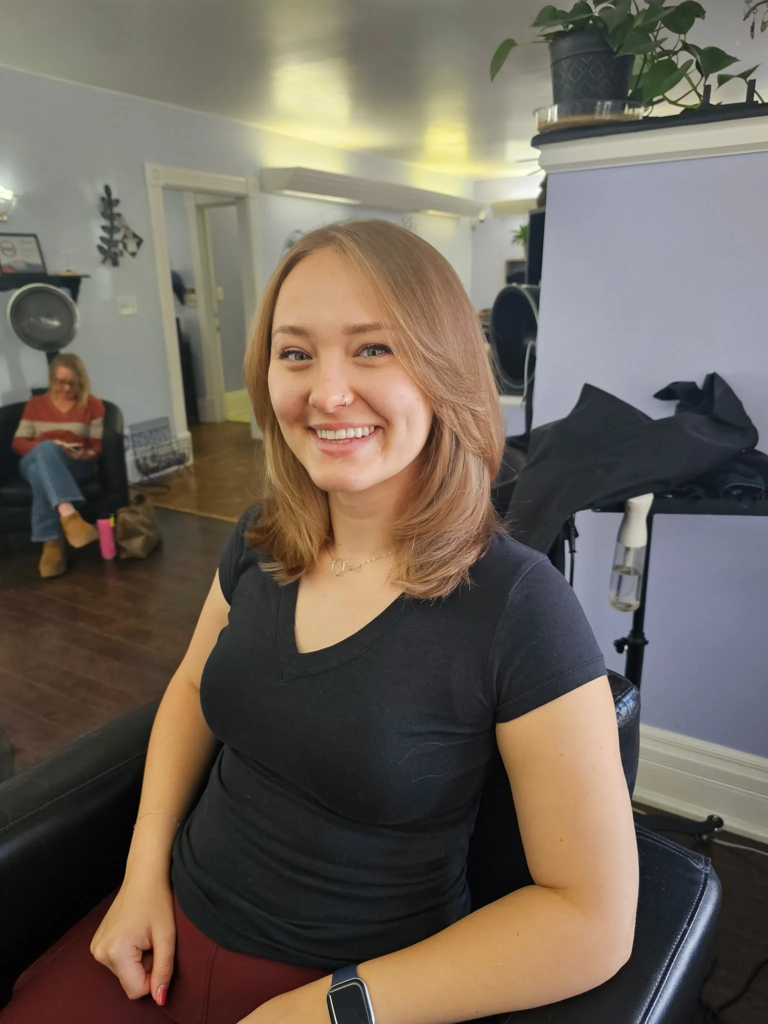 Young woman with shoulder-length light brown hair smiling, sitting in a salon chair, wearing a black t-shirt, gold necklace, and smartwatch. Hair salon with hair drying machine and another woman in the background. Radiant Styles by Julie