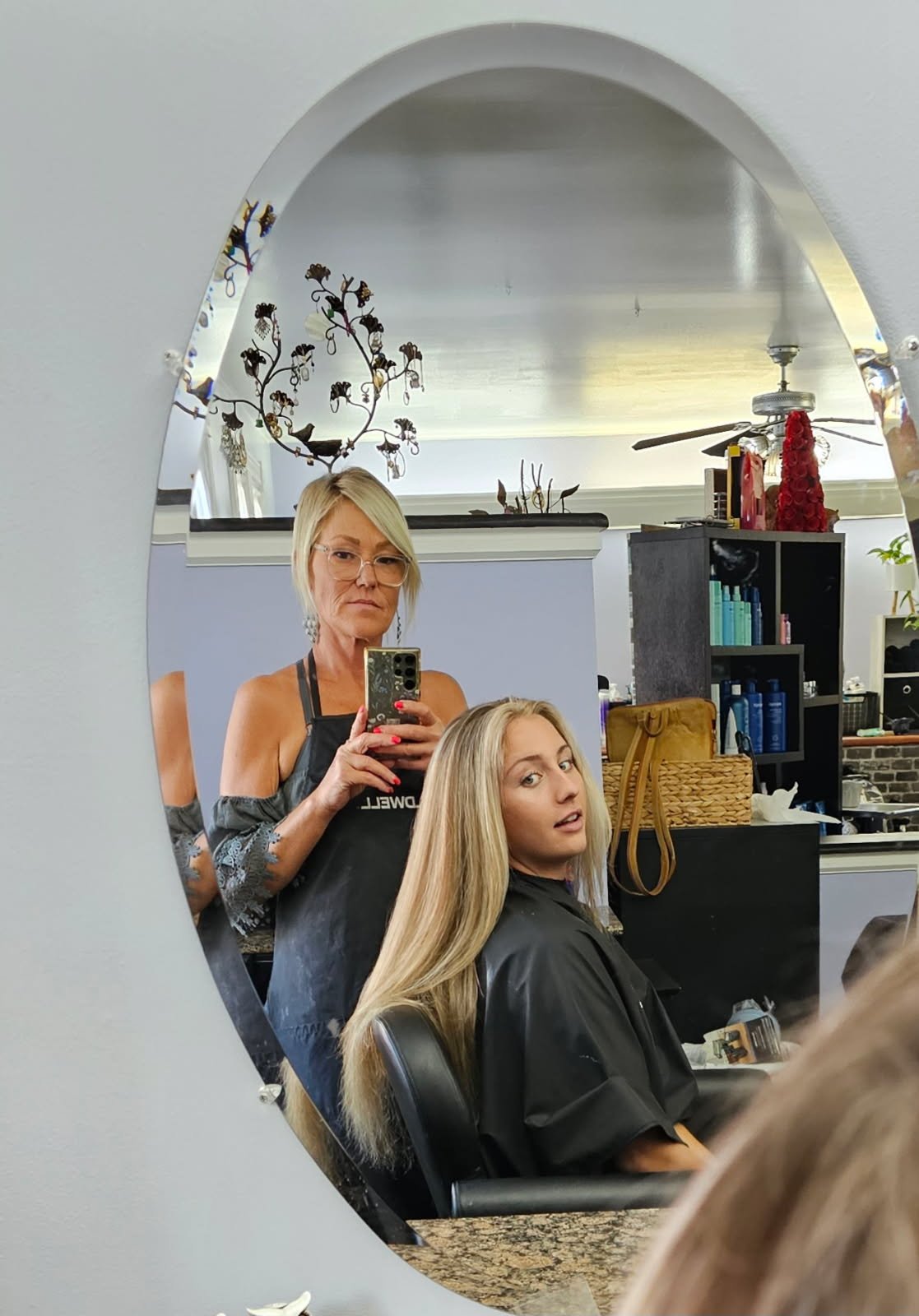 A woman with long blonde hair sitting in a salon chair with a stylist taking a mirror selfie. The stylist has short blonde hair, glasses, and is wearing a black apron. The salon background includes shelves with hair products, a basket, and decorative