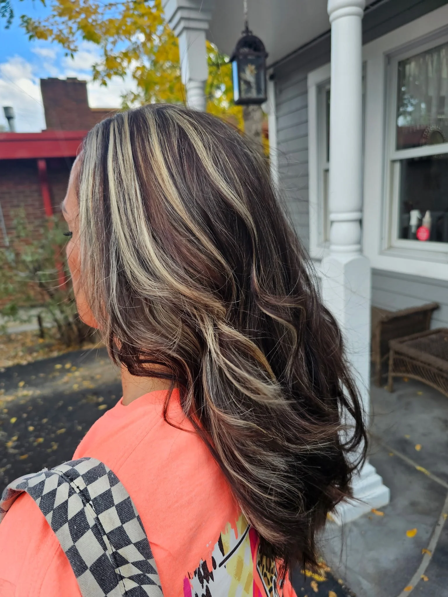 Side view of a woman with long wavy hair with blonde highlights, wearing an orange top with a black and white checkered bag strap, standing outside in front of a house with a porch, surrounded by fallen autumn leaves. Radiant Styles by Julie