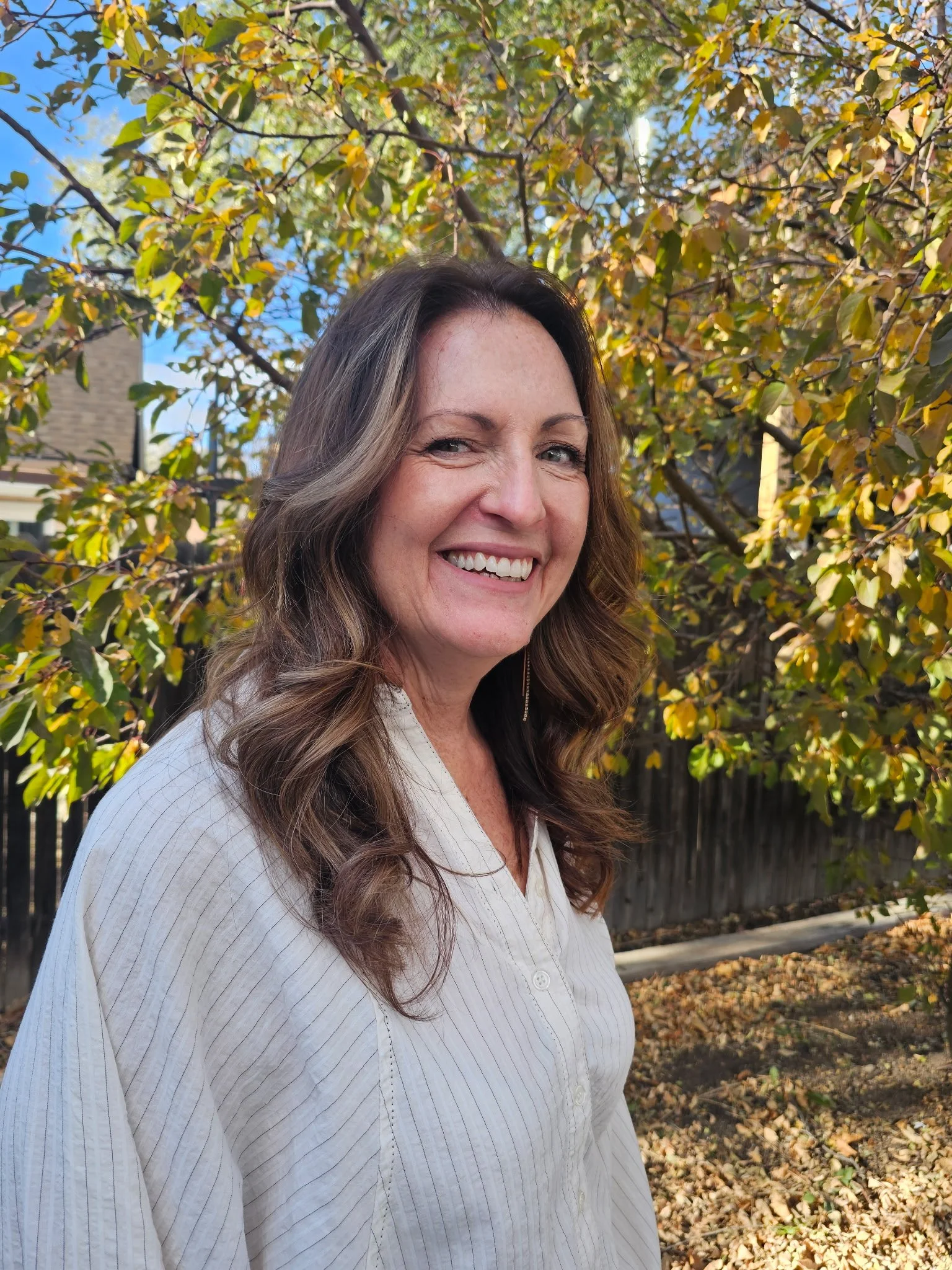 A woman smiling outdoors during autumn, standing in front of a tree with yellow and green leaves, with a residential neighborhood and blue sky in the background. Radiant Styles by Julie