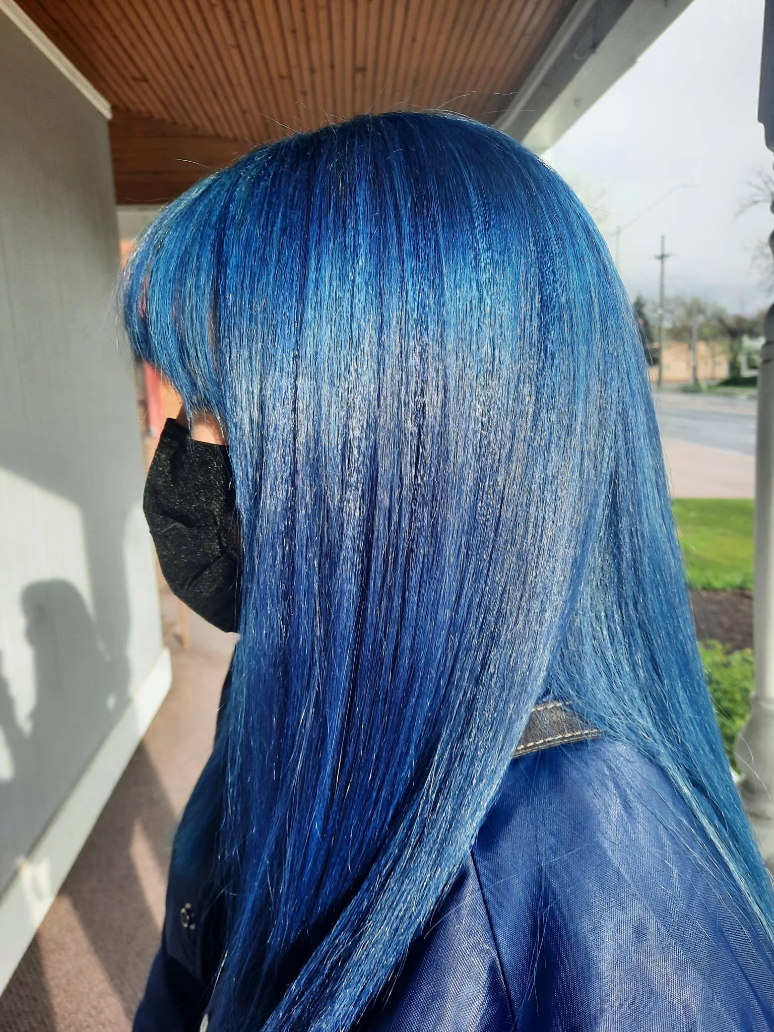 Side view of a person with long, straight, bright blue hair, wearing a black face mask and a blue jacket, standing outdoors near a building with a porch and a grassy yard. Radiant Styles by Julie