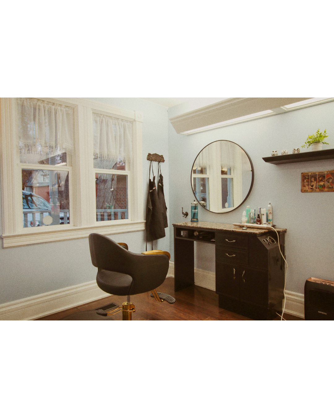 A vintage barber or hairstylist station with a black chair, a mirror, and a granite-topped black cabinet, in a room with white walls, large windows with lace curtains, and a small black shelf with decor and plants.