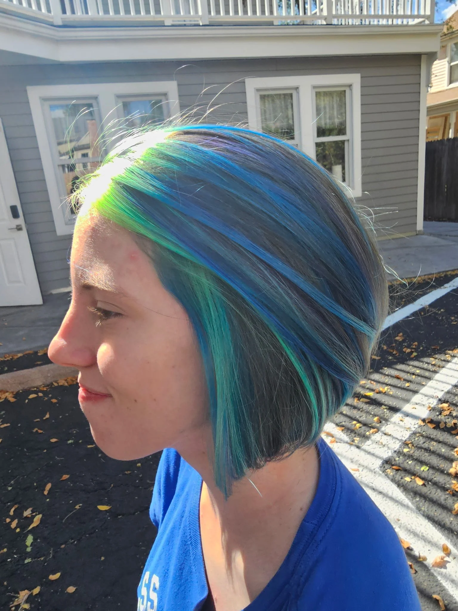 Side profile of a person with short, straight hair dyed in shades of blue, green, and blonde, standing outdoors near a gray house with white-framed windows, during daylight. Radiant Styles by Julie