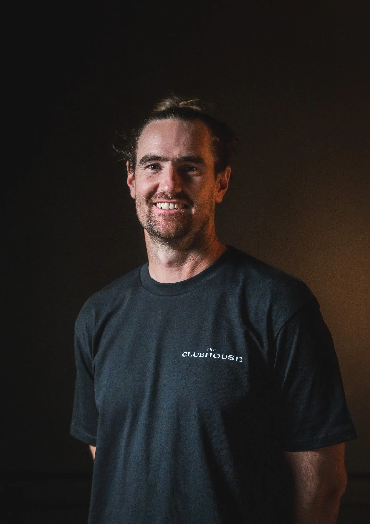 A smiling man with dark hair tied back, wearing a black T-shirt with 'The Clubhouse' logo on it, standing against a dark background.