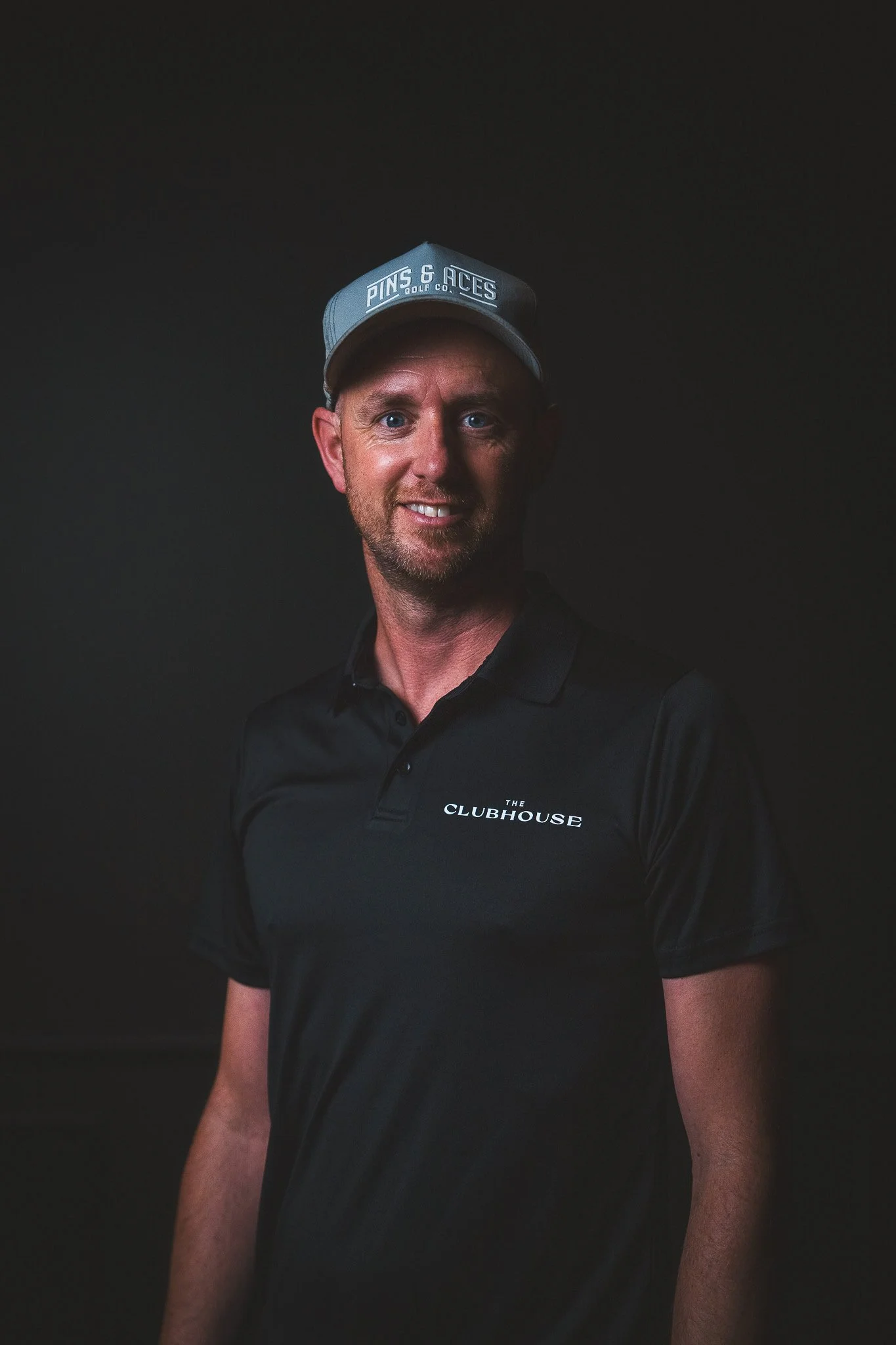 A smiling man wearing a black polo shirt with the words "The Clubhouse" and a black cap with the words "Pins & Aces Golf Co." stands against a dark background.