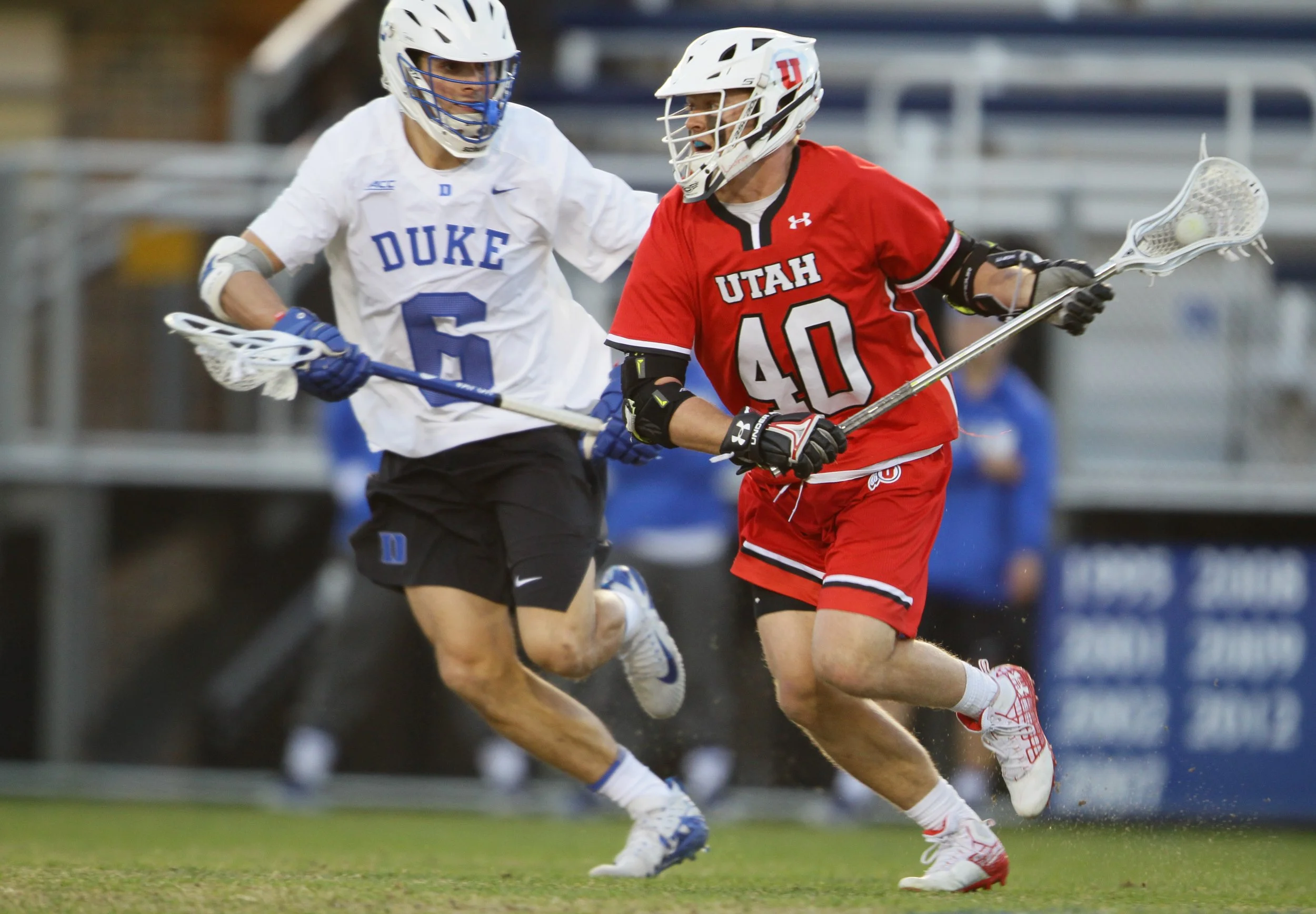 Lacrosse players from Duke and Utah competing on a field, with Duke player in white and black uniform (#5) and Utah player in red uniform (#40), both wearing helmets and gloves, fighting for possession of the ball.